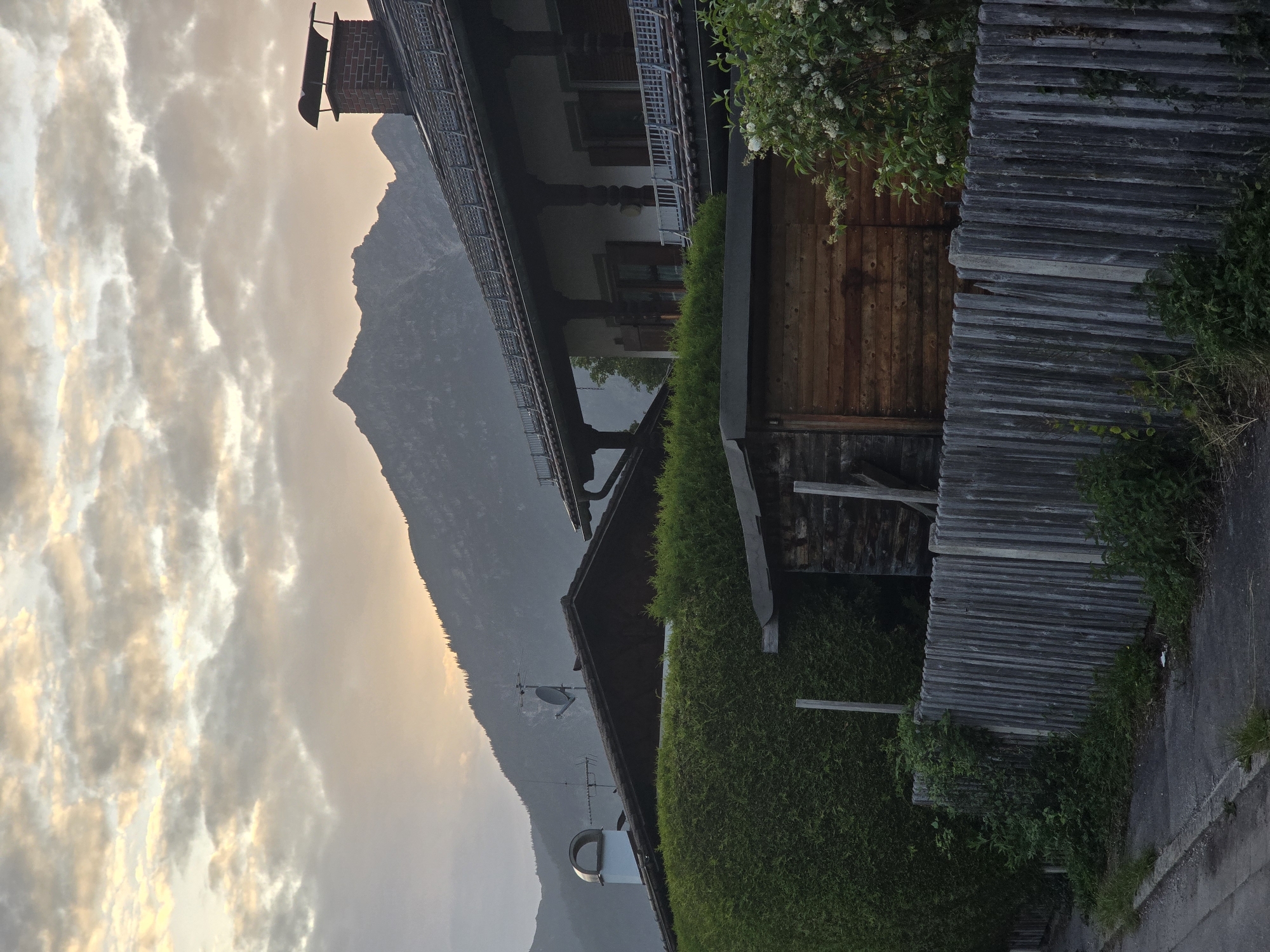 The image depicts a view of a suburban area in Garmisch-Partenkirchen, Germany, at dusk. In the foreground, a grey, weathered wooden fence runs horizontally across the frame. Beyond the fence, a neatly trimmed green hedge is visible, with a small wooden shed situated behind it. Further back, parts of two residential buildings are visible. The building on the right has a dark tiled roof with a brick chimney and dormer windows. The building on the left has a dark roof with various antennas and a white chimney. In the background, a large, dark mountain range dominates the skyline, with its peak silhouetted against a sky filled with soft, cloudy, twilight hues of orange and grey. The overall impression is one of quiet residential life set against a dramatic natural landscape.
