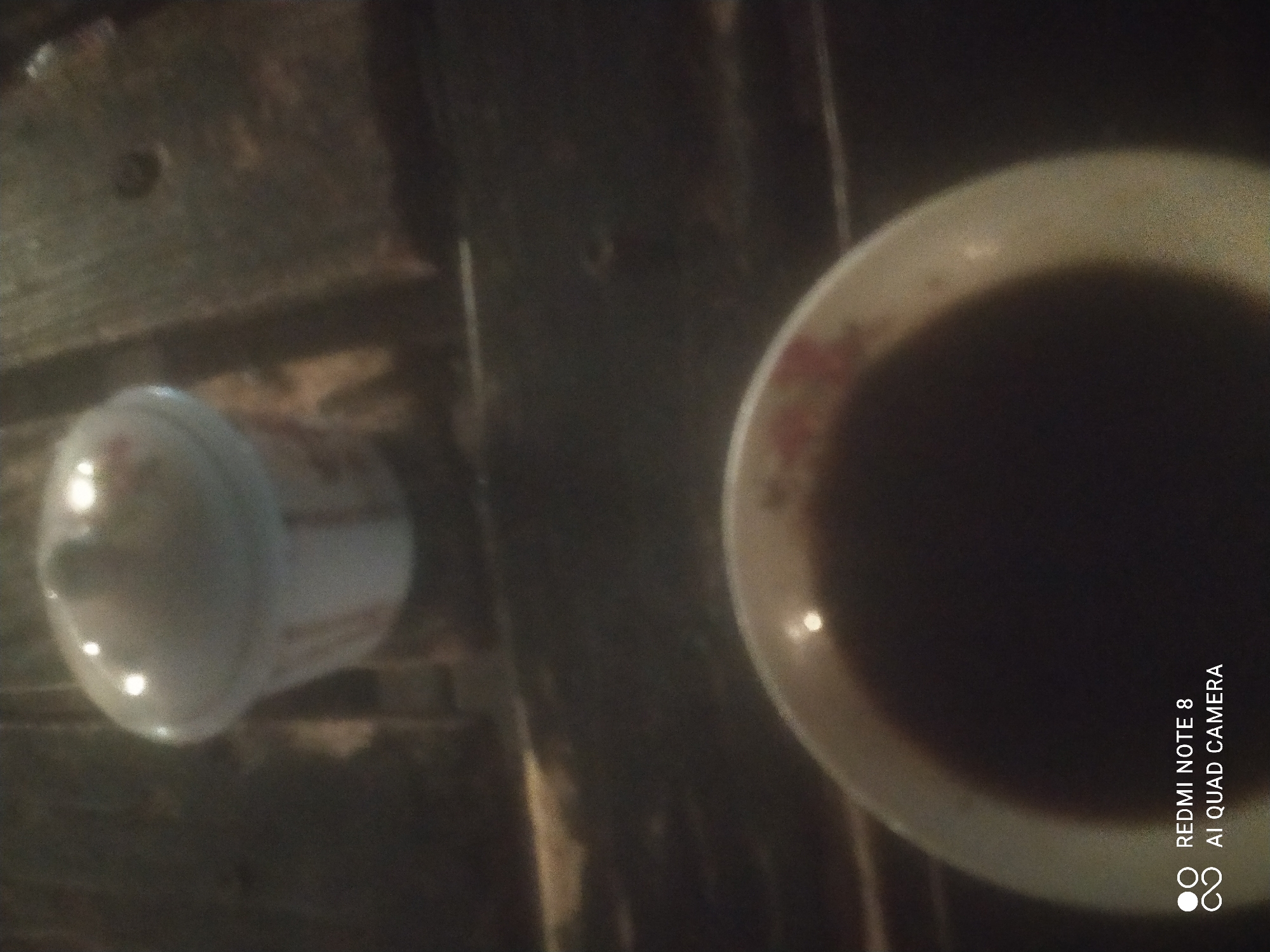 The image shows a close-up, dimly lit shot of two ceramic containers on a wooden surface. The upper container appears to be a lidded dish, possibly a sugar bowl or small pot, with a floral pattern. Below it, a shallow bowl filled with a dark liquid, likely coffee or tea, is visible. The liquid's surface reflects light. The wooden surface has a rough, planked texture. The bottom left corner of the image displays a watermark with "REDMI NOTE 8" and "AI QUAD CAMERA". The overall lighting is poor, suggesting the photograph was taken in low-light conditions, possibly at night. The image does not contain any elements that indicate a specific location in Sumurber, Indonesia.