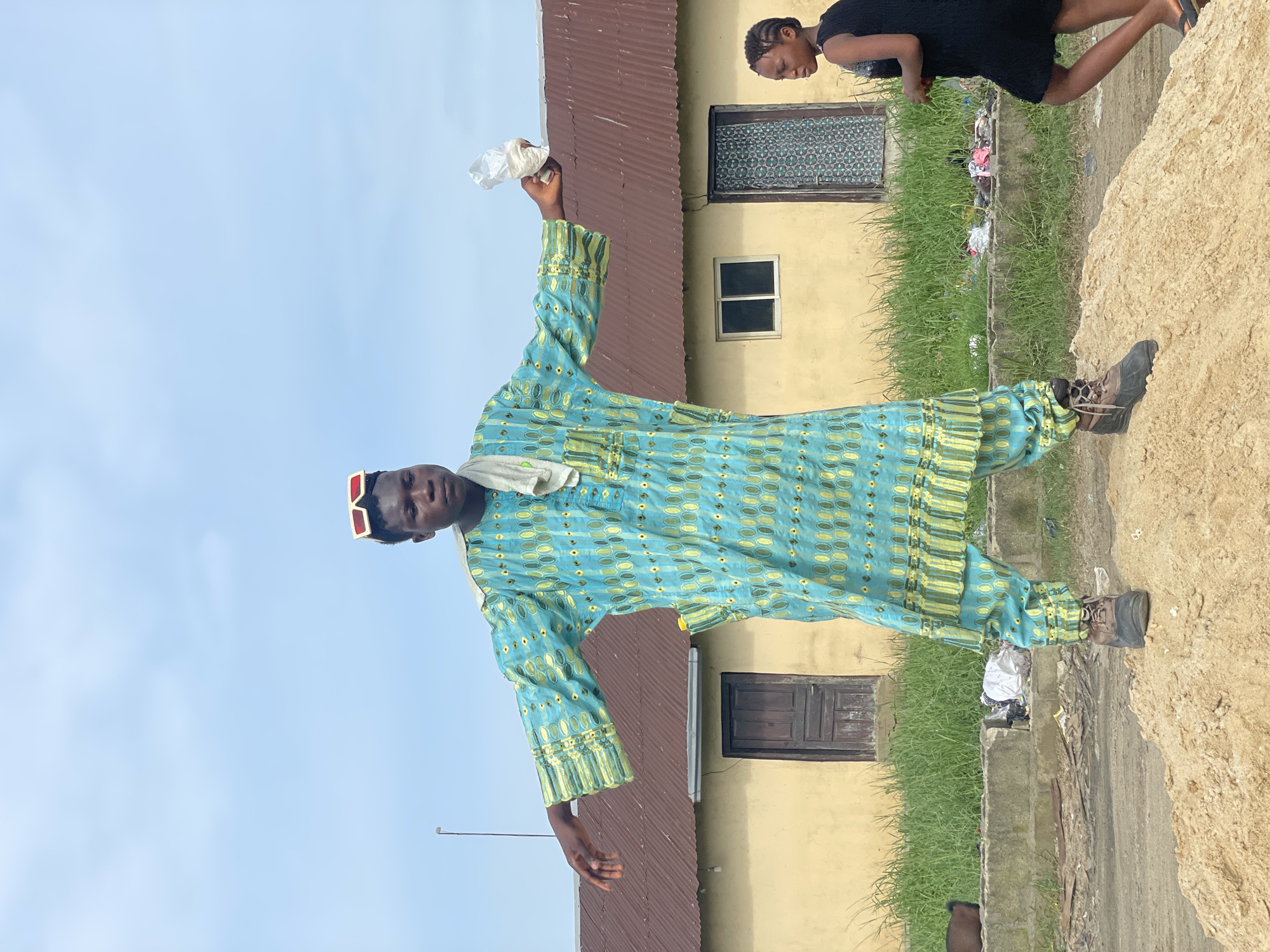 A man stands outdoors, wearing a blue and yellow patterned robe, brown boots, and red sunglasses, and holding a crumpled white object in his right hand. Behind him is a yellow building with a brown roof and two dark windows. A woman in a black dress stands partially visible at the upper right corner of the image. There is a pile of light-colored sand or earth in the foreground and grass growing along the base of the building. Some scattered trash is also visible. The sky is a light blue color. This scene takes place in the city of Ado, Nigeria.