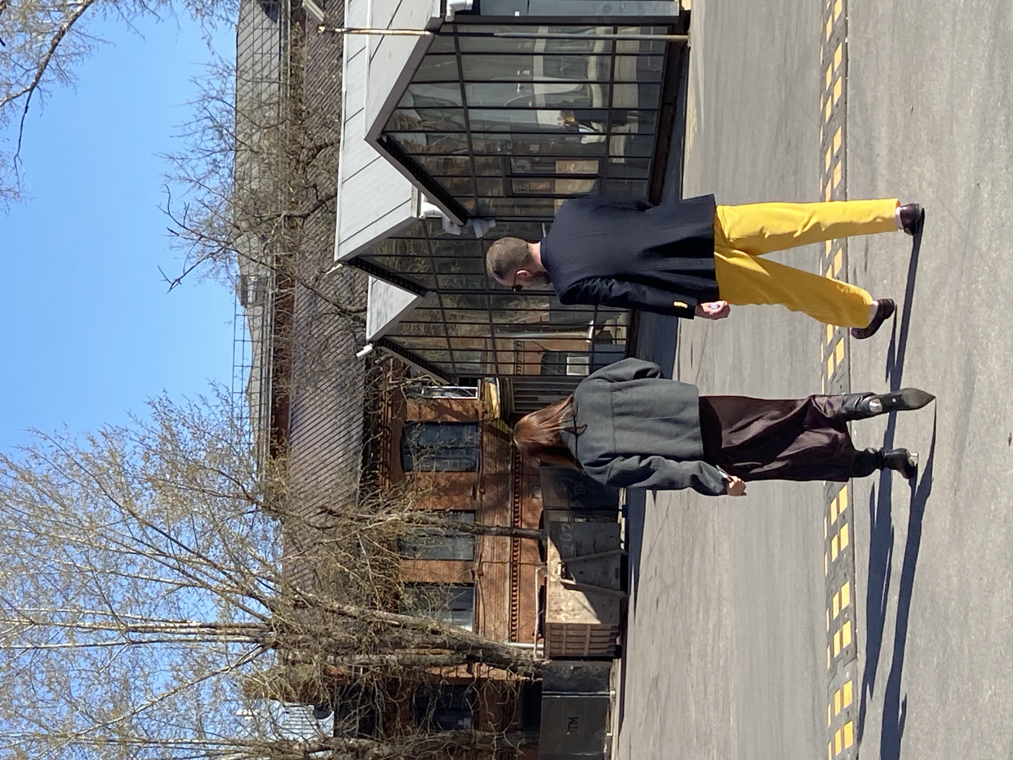 The image captures two individuals walking on a sidewalk next to a building in Moscow, Russia. One person is wearing a dark blazer, yellow pants, and sunglasses. The second individual is wearing a dark overcoat and dark pants. The backdrop includes an industrial-style building with glass windows. The ground has yellow markings. A bare tree is on the left side of the image.