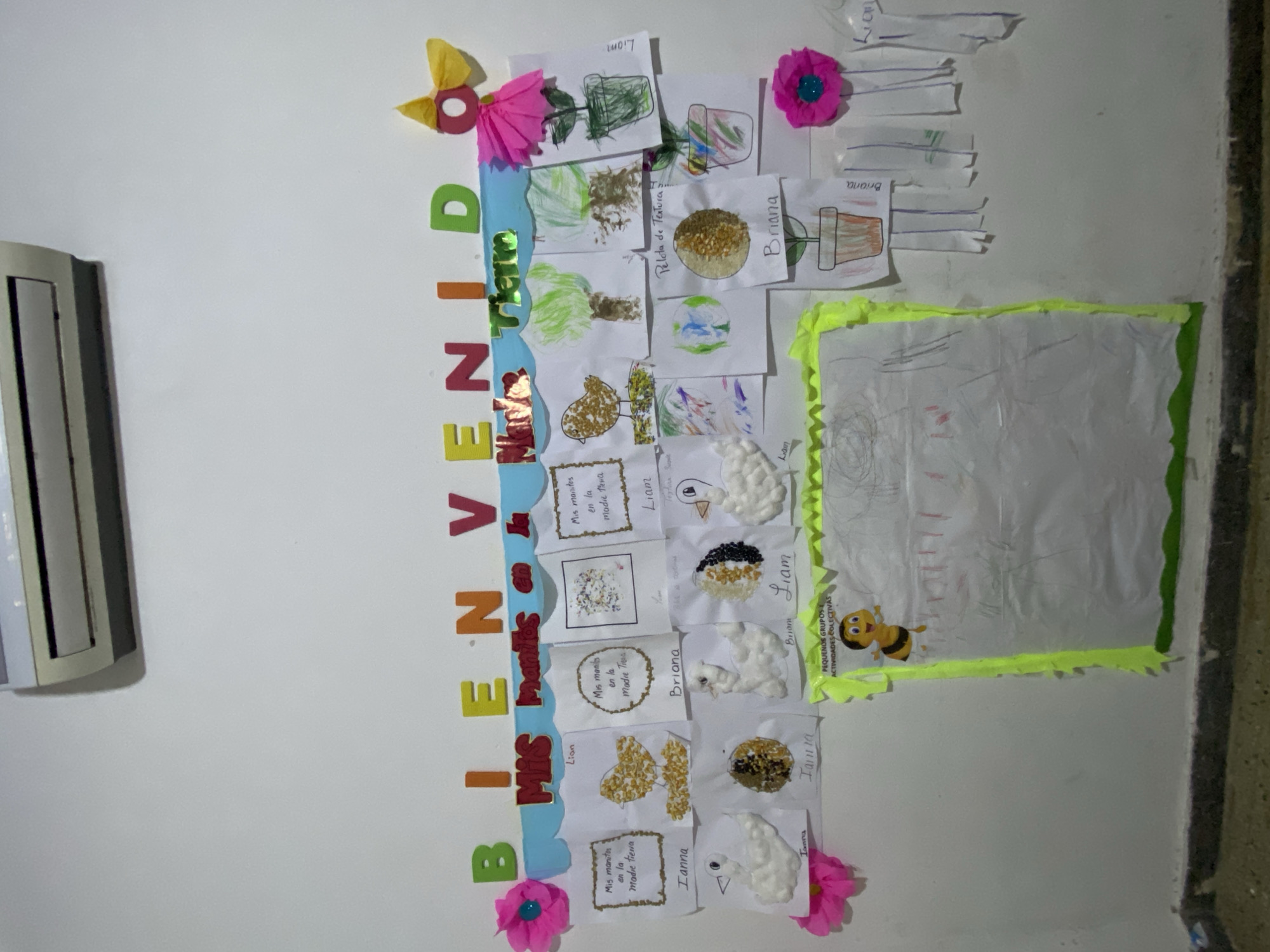 The image shows a vertically oriented display mounted on a light-colored wall. The display spells out "BIENVENIDO" with colorful block letters on a light blue backdrop, decorated with paper flowers. Below this greeting, there are several children's artworks, likely drawings or craft projects, attached to the board. These artworks appear to have the names of the children, "Ianna", "Briana", and "Liam", written on them. The artworks consist of drawings and craft projects. There's a large square frame with a yellow border, possibly containing more artwork or information. A wall-mounted air conditioner unit is visible on the left side of the image. The location in the city of Tinaquillo, Venezuela is not supported by the information in the image.
