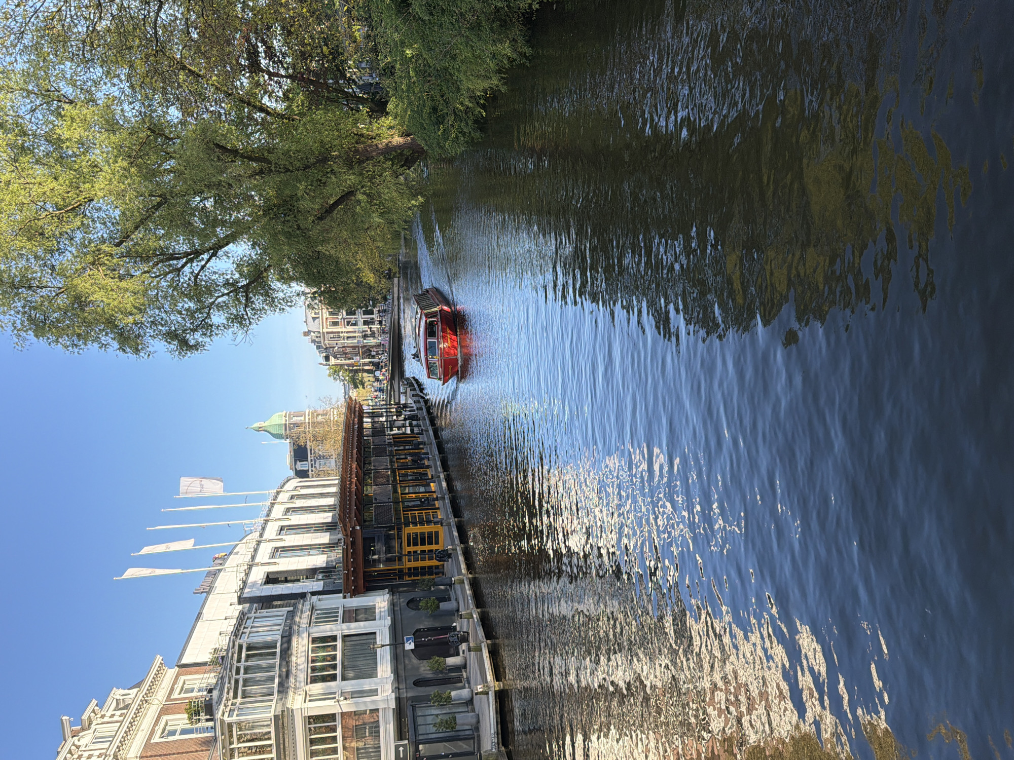 A serene urban canal scene in Amsterdam, Netherlands, unfolds under a clear blue sky on a bright, sunny day. The image is taken from a slightly elevated perspective, looking down the length of the canal.
A vibrant red canal boat, appearing to be a tour boat, glides through the calm water in the middle distance, leaving a subtle wake behind it. The water reflects the deep blue of the sky and the lush green of the surrounding trees, creating shimmering patterns of light and shadow.
On the left bank (from the viewer's perspective, after proper orientation), a dense canopy of mature green trees overhangs the water, adding a natural, verdant element to the cityscape. On the right bank, a series of architectural structures line the canal. In the foreground, a modern building with large glass windows and grey facades is visible. Further along the canal bank, a long, dark floating platform with distinctive yellow railing structures and shaded seating suggests a waterside cafe or restaurant. Beyond this, traditional Amsterdam buildings with gabled roofs and brickwork rise, featuring multiple windows. A prominent building with a distinctive green dome is visible in the distance. Several white flags fly from poles attached to one of the buildings; however, any text on them is too distant to be legible. A small directional sign with an arrow pointing left is visible on a lower structure near the water, but its text is also illegible.
There are no people distinctly visible in the frame. The overall impression is one of peaceful urban activity on a pleasant day.