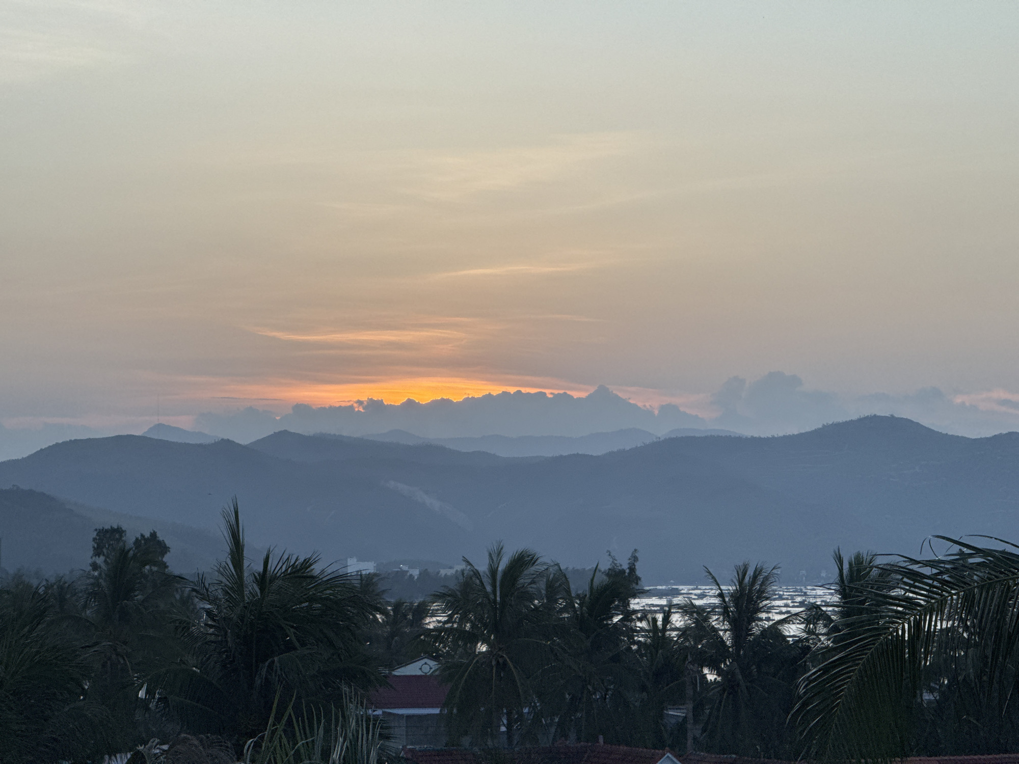 The image shows a serene sunset scene over the city of Song Cau, Vietnam. The sky is a gradient of soft oranges, pinks, and yellows, transitioning into a pale blue at the top. In the foreground, silhouetted palm trees with their fronds spread out create a natural frame for the landscape. Beyond the trees, layers of hazy, blue mountains recede into the distance. A body of water, likely a bay or lagoon, is visible between the mountains and the foreground, reflecting the colors of the sky. Scattered buildings and what appear to be small boats or structures are present on the water. The overall atmosphere is peaceful and tranquil.
