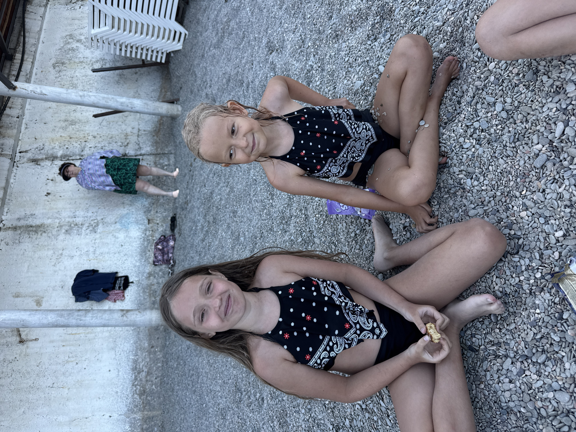 The image shows two young girls and an adult sitting on a pebble beach. The girls are wearing swimsuits and appear to be enjoying themselves. One girl is looking at the camera and smiling, while the other is looking down at a piece of food she is holding. The adult is wearing a checkered shirt and a green skirt and is standing in the background. The beach is covered in gray pebbles, and there are some white lounge chairs stacked in the upper left corner of the image. The setting appears to be a sunny day at the beach, possibly in Partenit, Ukraine, given the location context. There is no visible text in the image. The overall mood is relaxed and casual, suggesting a day of leisure and enjoyment.