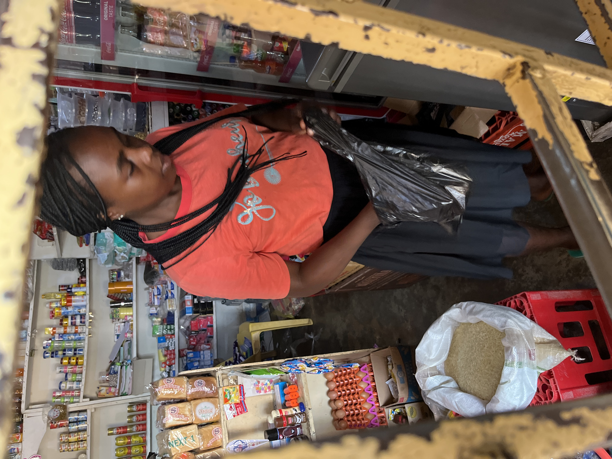 The image captures a young woman with braided hair standing in what appears to be a small convenience store or market stall in Bukuru, Nigeria. She is wearing an orange t-shirt with a light blue graphic and a dark skirt, and is holding a black plastic bag. Shelves behind her are stocked with various food items, canned goods, and other products. A refrigerator with soft drinks is visible to her right. In the foreground, a large bag of rice is placed on a red crate, with the contents spilling out. The scene is framed by a yellow, weathered metal structure, suggesting an outdoor or semi-enclosed retail space. The lighting is somewhat dim, casting shadows.