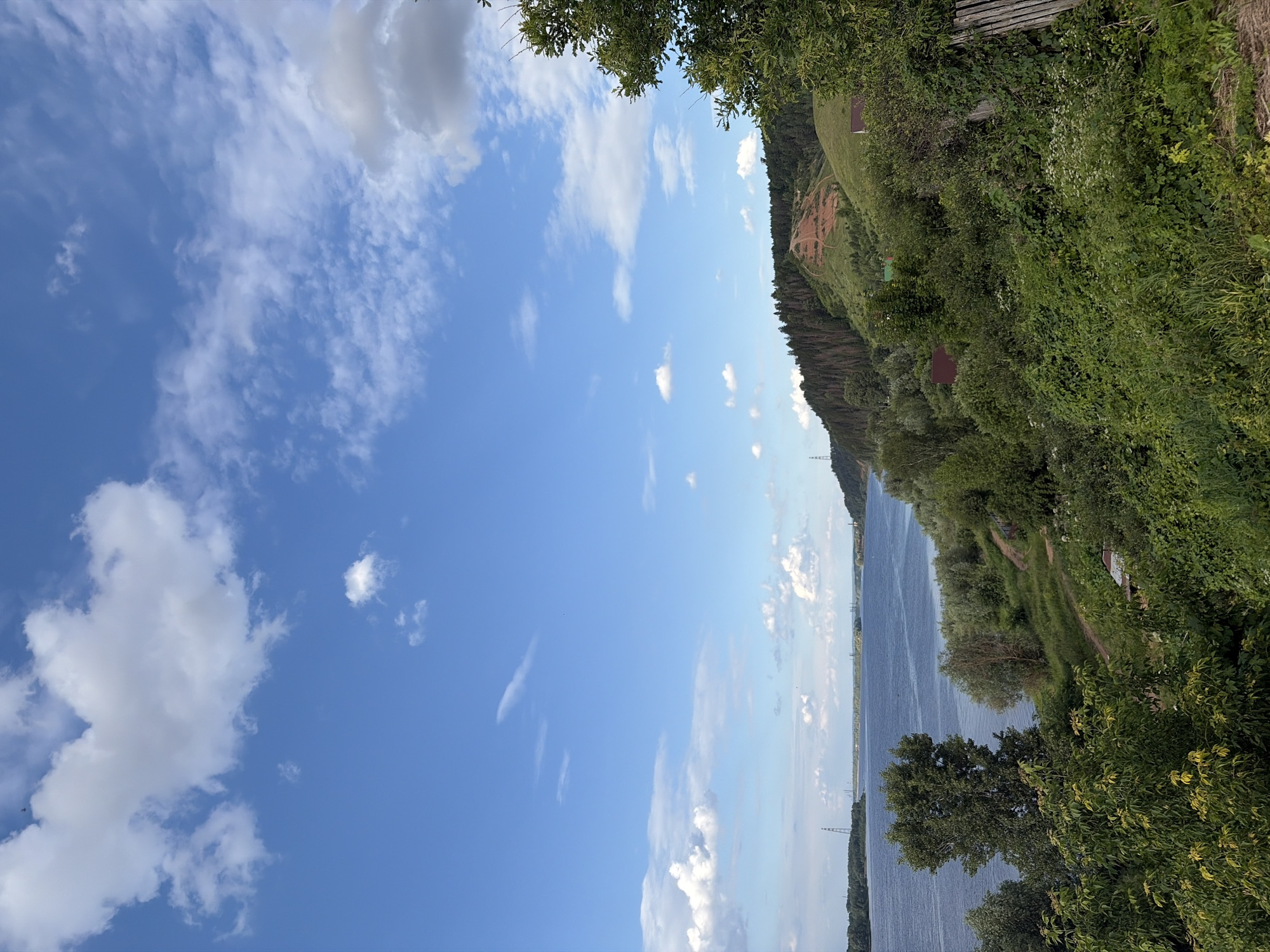 The image depicts a scenic riverside landscape, likely taken in Olkhovka, Russia. The scene is dominated by a wide, calm river that stretches into the distance, its surface reflecting the bright blue sky and scattered white clouds. The left side of the frame is filled with this expansive sky, featuring wispy cirrus clouds and larger, fluffier cumulus formations.
The right side of the image shows a lush, green embankment rising steeply from the river. Dense vegetation, including trees and bushes, covers the hillside, creating a vibrant tapestry of green. A narrow, winding path is visible on the embankment, suggesting a hiking or walking trail. Several small, reddish-brown structures, possibly cabins or sheds, are nestled amongst the greenery. In the distance, a tall power line pylon can be seen against the horizon.
The time of day appears to be late morning or early afternoon, indicated by the bright sunlight and clear blue sky, although some shadows suggest the sun may be starting its descent. The weather is fair and pleasant, with only a few clouds dotting the sky. There is no visible text in the image. The overall mood is tranquil and serene, inviting contemplation of nature's beauty.