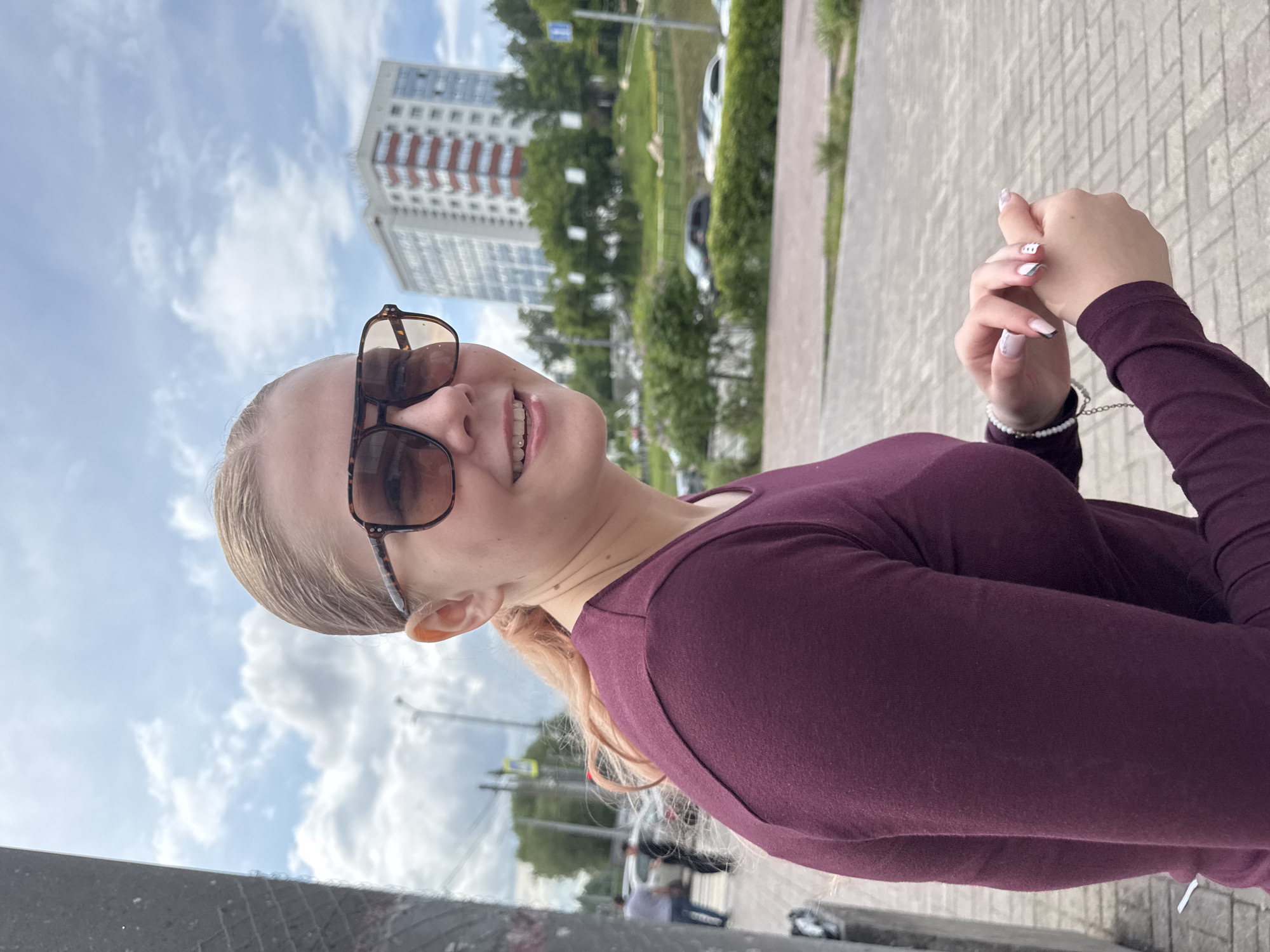 A young woman with light-colored hair, wearing dark sunglasses and a maroon long-sleeved top, is captured in a casual outdoor portrait. She is smiling faintly towards the camera, revealing braces on her upper teeth and a small stud on her lower lip. Her hands are clasped together in front of her chest, showcasing white nails with abstract black and gray patterns, and a white beaded bracelet on her left wrist.
The setting appears to be an urban environment during the daytime, under a bright, partly cloudy sky. To her left, a tall, multi-story residential building with light-colored walls and red accents is visible in the background. To her right, beyond the building, are green trees, a paved road with parked or moving vehicles, and a glimpse of other distant structures. The foreground consists of a textured, light-colored paved surface, likely a sidewalk. The overall mood is relaxed and pleasant. No discernible text is visible in the image.