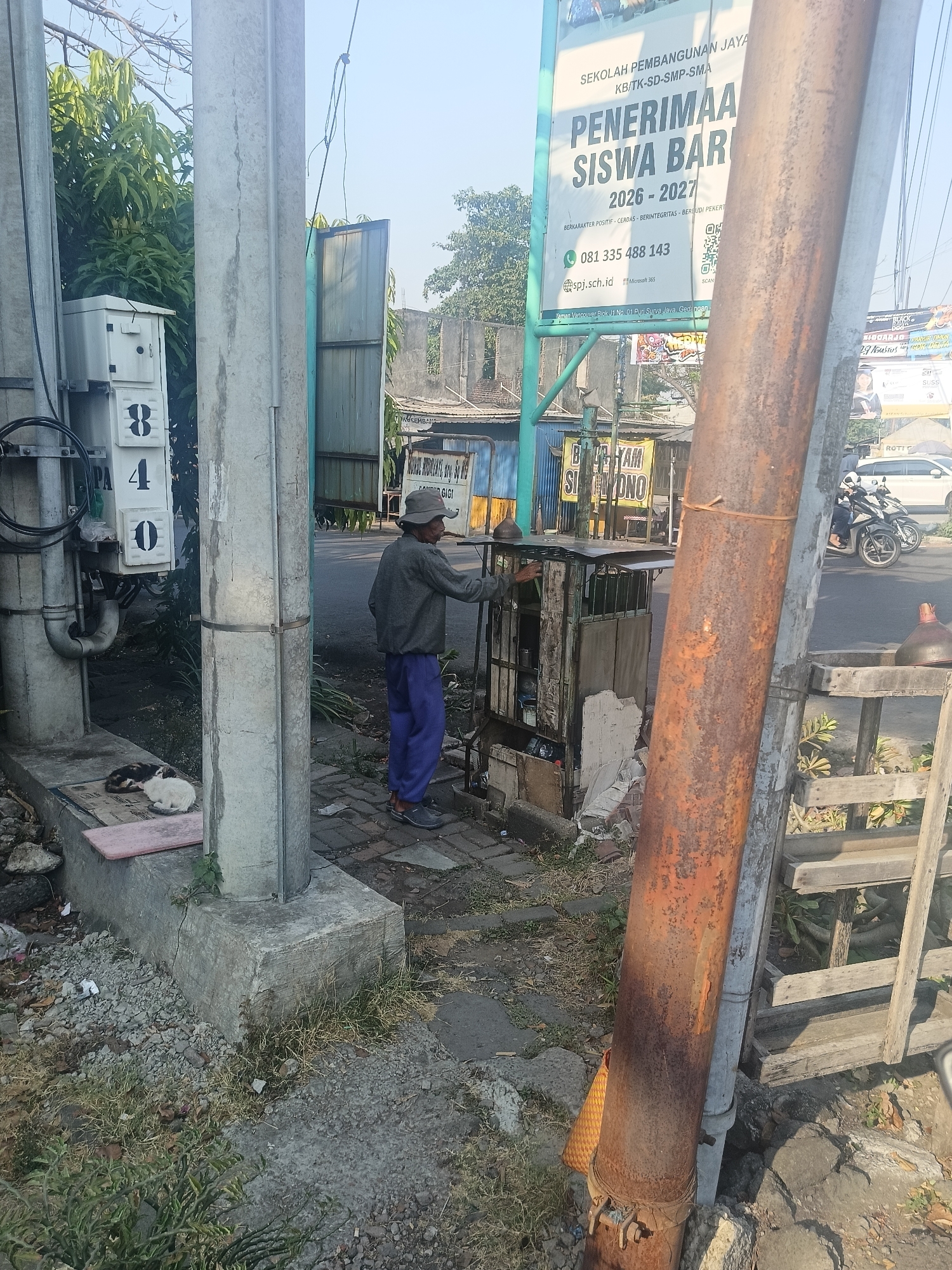 In Wedi, Indonesia, a man wearing a grey long-sleeved shirt and blue pants, along with a wide-brimmed hat, stands beside a small, weathered cart. He appears to be interacting with something on the cart. Nearby, two cats are resting on a concrete ledge next to a utility box with the numbers 8, 4, and 0 displayed on it. A large, rusty pipe stands prominently in the foreground, parallel to a concrete utility pole. In the background, a billboard advertises the "SEKOLAH PEMBANGUNAN JAYA," indicating an educational institution. The scene unfolds on a street with vehicles, including motorcycles and a white car, visible in the distance. The environment suggests an outdoor setting with buildings and trees.