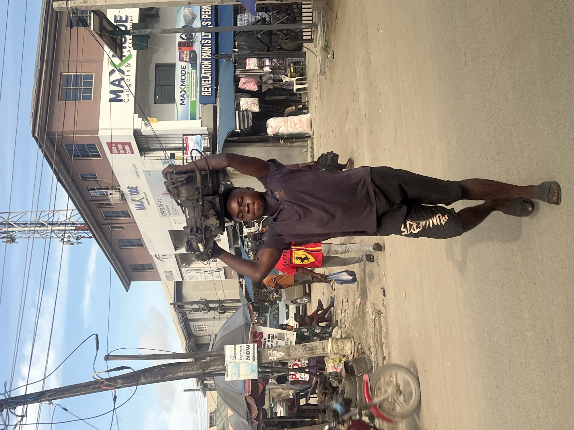 The image captures a vibrant street scene in Lagos, Nigeria, during daytime under a clear blue sky. The primary subject is a man, dressed in a dark t-shirt and black shorts, confidently carrying a heavy, dark-colored mechanical object, possibly an engine or generator part, balanced on his head. He looks directly at the camera with a composed expression.
The background features a bustling urban environment. On the left, a multi-story building prominently displays "MAX MODE Gadgets & Accessories" signage on its brown and white facade. Other visible text on this building includes "Service Centre," "CLEVELAND," and "OPPO A6 Pro." To the right, another commercial building has a sign for "REVELATION PAINTS LTS." The street is active with other pedestrians; one person is visible wearing a red shirt with a Ferrari logo, and others are seen near street stalls. A utility pole with numerous crisscrossing power lines is visible in the foreground, and hints of parked vehicles and motorcycles can be seen throughout the busy street. Further signs include "DON'T WAIT, START NOW" and partially visible text like "ARGES or N100" and "Cor #200." The man's shorts also bear the text "PUNGERS." The scene portrays everyday life and commerce in a lively urban setting.