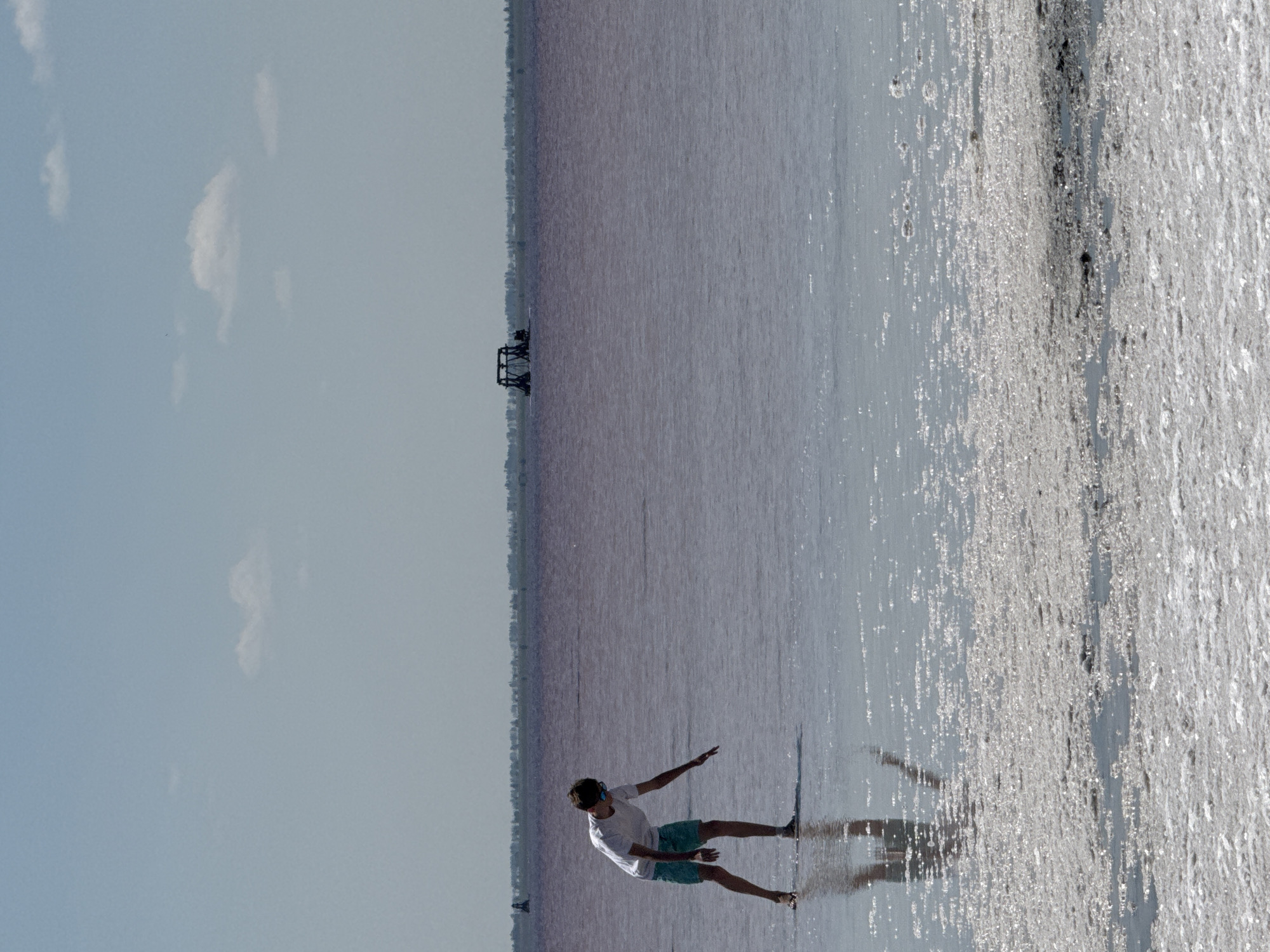 The image depicts a boy standing in shallow, reddish-pink water with a salt-encrusted shore in the foreground. The boy, wearing a white t-shirt and blue shorts, is bending over, reaching his hand towards the water. The water appears shallow, with his legs and reflections visible. In the background, a thin line of trees or buildings is visible on the horizon, and a dark, industrial-looking structure, possibly part of a salt-harvesting operation, is present on the water further in the distance. The sky is a pale blue with a few scattered clouds. This scene is consistent with the environment of the salt lakes in Bursol', Russia, known for their unique coloration due to microorganisms and high salt content.