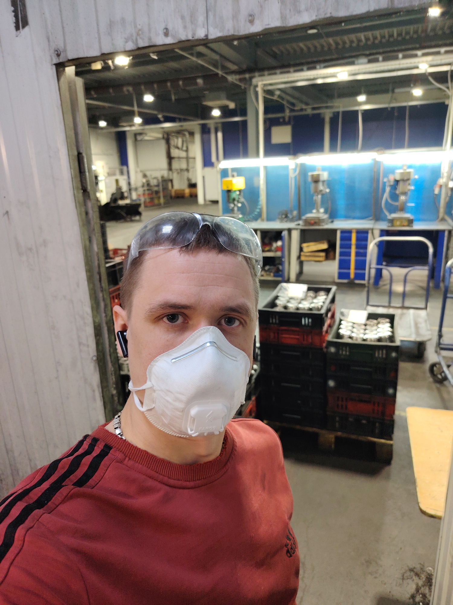 The image shows a person wearing safety glasses and a white face mask standing in what appears to be a factory or industrial setting. The person is wearing a red long-sleeved shirt with black stripes on the shoulders. Behind the person are stacked crates filled with metallic items, and further in the background is a workbench with drilling machines. The location is presumed to be a workshop in Noviy, Russia, based on the user’s prompt.