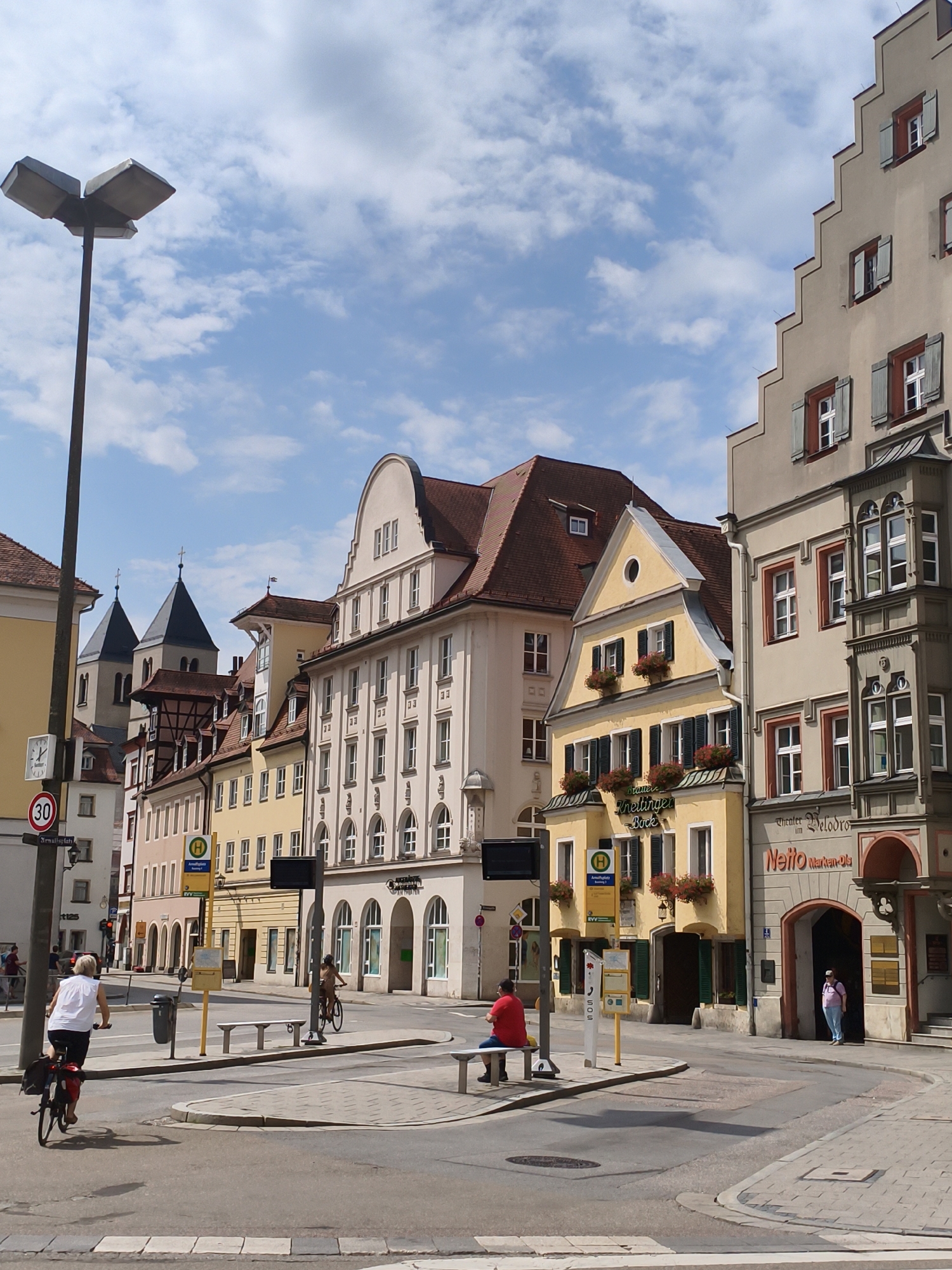 The image captures a street scene in Regensburg, Germany, likely in the historic city center. The architecture features a mix of old and new buildings, with a prominent yellow building housing "Bäckerei Krettinger" and "Theater Velodrom." A "Netto Marken-Discount" grocery store is visible with an arched entrance. Several people are present: a woman on a bicycle in the foreground, another person cycling in the background, and individuals near the entrance of the store. Traffic signals and street signs, including a "30" speed limit sign, are visible. The sky is partly cloudy, suggesting a daytime setting. The overall impression is a lively urban environment with a blend of commerce and daily life.