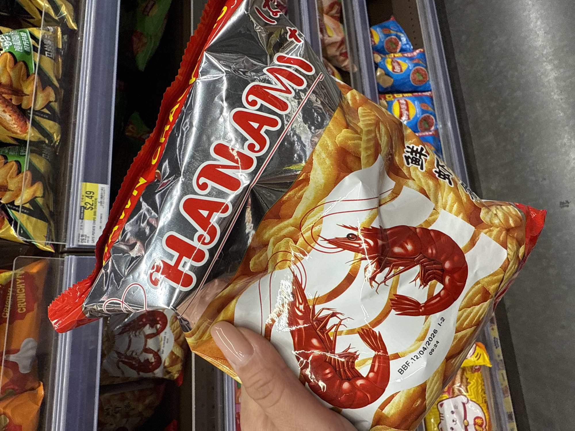 A hand holds a bag of Hanami brand prawn crackers in a store, likely a supermarket, with shelves of other snack foods visible in the background. The packaging is predominantly red, silver, and white, featuring images of prawns and what appears to be crunchy chips. The Best Before date on the bag reads "BBF.12/04/2026". A price tag showing "$2.49" can be seen on a nearby shelf. The location is likely Orlando, United States, based on the prompt.