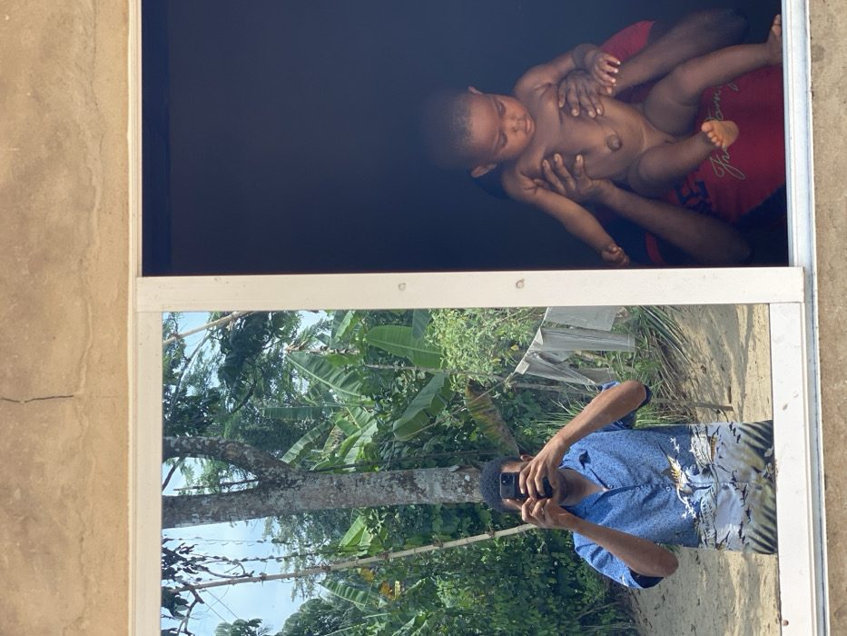 In Ikot Ete Ekpo, Nigeria, the image presents a bifurcated scene observed through a white-framed window. The upper section reveals a light brown, rough-textured concrete wall with minor cracks.
The left pane of the window functions as a reflective surface, showing the outdoor environment and the photographer. A dark-skinned adult, presumably male, is reflected from the chest up, holding a dark smartphone directed towards the scene, thus capturing their own image. He wears a short-sleeved blue collared shirt with a light-colored graphic depicting ocean waves and a fish. Behind the reflected individual, a lush, tropical outdoor setting is visible, characterized by prominent green foliage, large-leafed plants resembling banana trees, tree trunks, and a sandy or dusty ground. Light-colored laundry is seen hanging on a line or bush in the background.
The right pane of the window offers a direct view into a dark, interior space. A dark-skinned infant, appearing unclothed, is held upright by hands belonging to an individual partially visible below. This individual wears a red short-sleeved t-shirt with a white graphic or text element. The infant looks downwards, and the interior background is dimly lit.