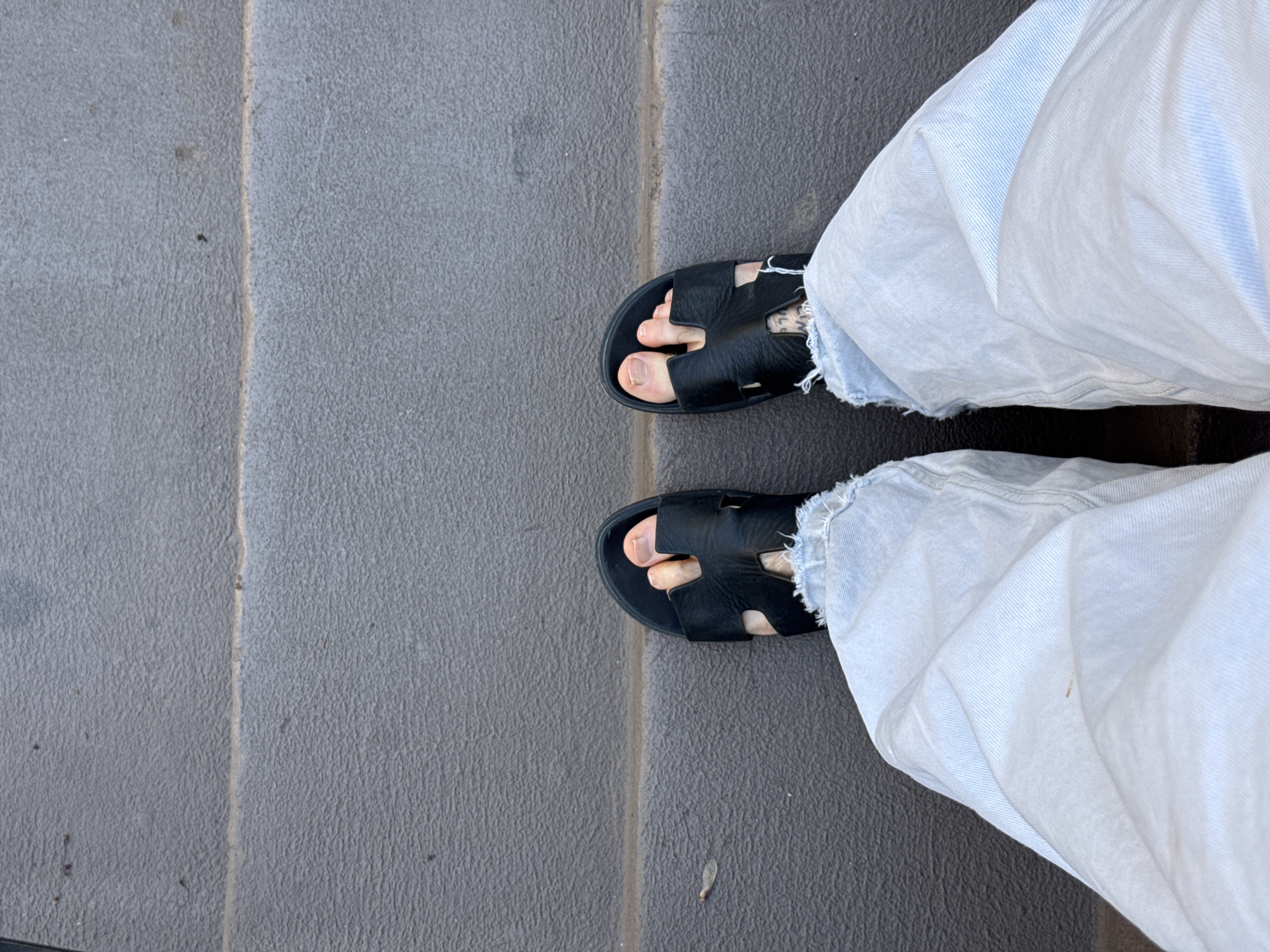 The image shows a view from above, looking down at a person's feet standing on a concrete staircase. The person is wearing light-wash denim jeans with frayed hems and black leather sandals. The staircase is made of gray concrete and appears to be outdoors. There is no information available in the image to verify if the location is in the city of Boston, United States of America.