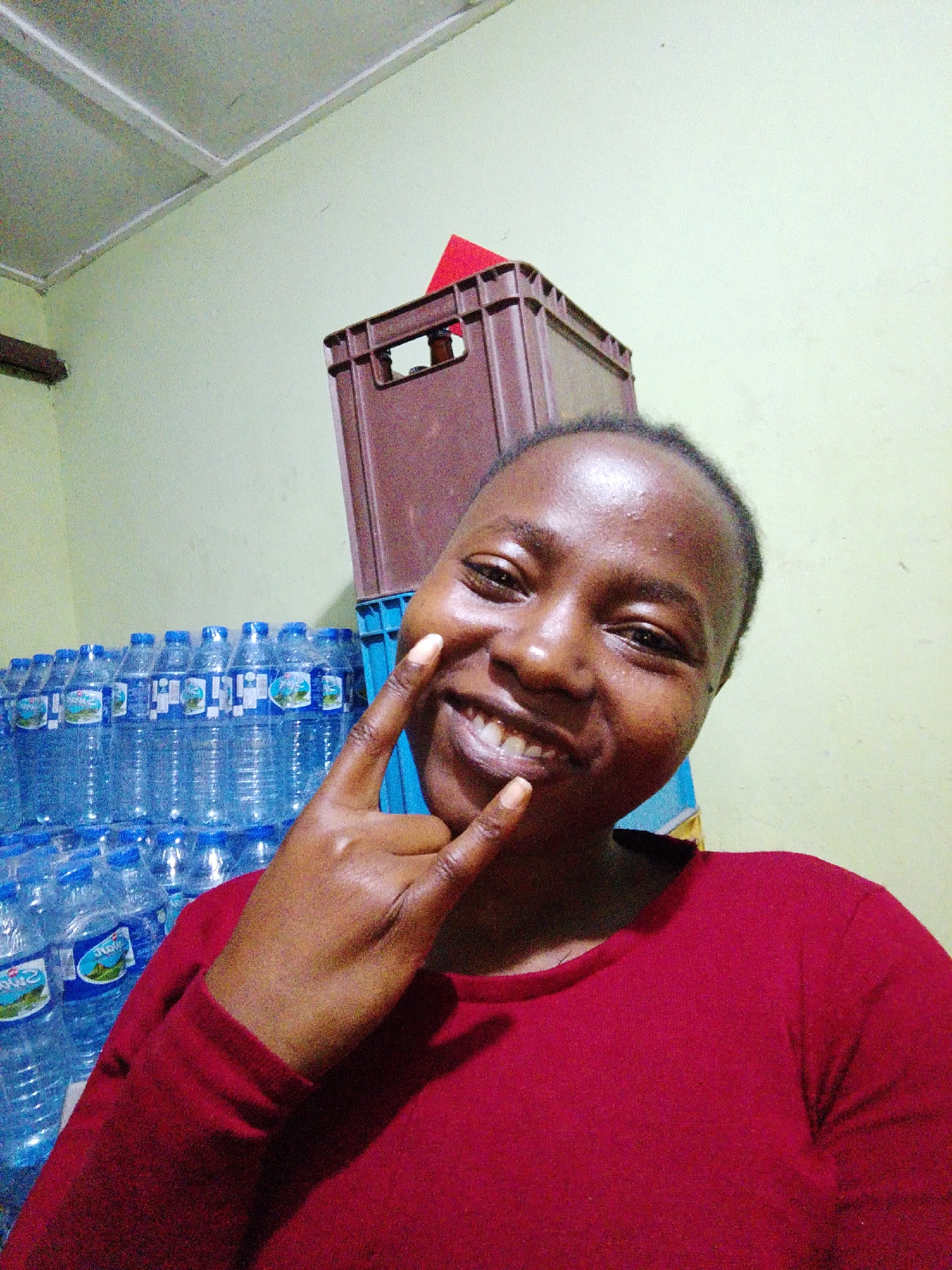 A person with dark skin and short dark hair, wearing a dark red long-sleeved shirt, smiles and makes a hand gesture resembling devil horns toward the viewer. The person is indoors, in front of a stack of bottled water on the left and stacks of plastic crates behind the water. The background includes a pale yellow-green wall and a partial view of a ceiling. The location is in the city of Unknown, Unknown.