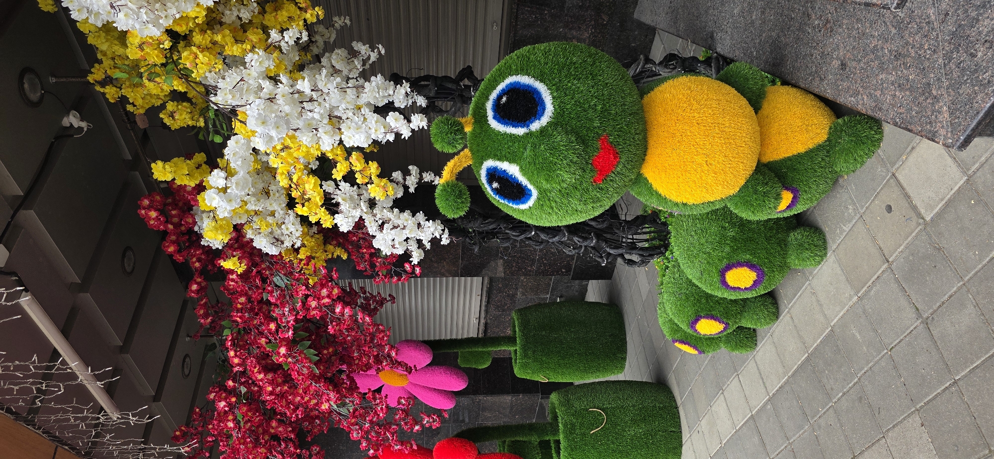 This image captures an outdoor decorative display in Kislovodsk, Russia, likely in front of a commercial establishment. The primary subject is a large, artificial green caterpillar sculpture with a yellow abdomen and large blue and white eyes. Its body is segmented, with each segment featuring a purple and yellow circular detail. The caterpillar is positioned on a paved sidewalk composed of gray square tiles.
Behind the caterpillar, a black, twisted metal structure supports an arrangement of artificial trees with branches laden with vibrant red, yellow, and white blossoms. To the left of the caterpillar, two additional artificial plants are visible, one resembling a large pink flower with an orange center, and another a red flower, both growing out of green, barrel-shaped bases covered in artificial grass.
The background features a building with a dark, textured facade and a closed, light-colored roll-up door. Above the display, a string of white LED icicle lights hangs from an overhang, casting a faint glow. The overall scene is static and appears to be a decorative arrangement for a business or public space, evoking a whimsical and colorful aesthetic. No people are present in the image.