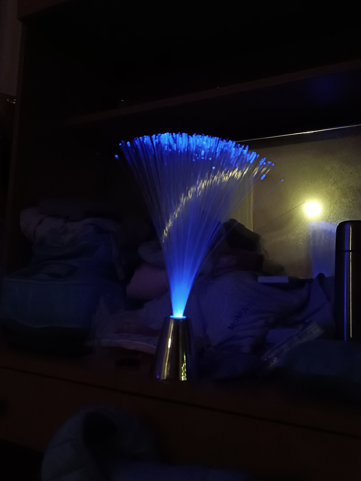 A fiber optic lamp with blue light is on a shelf or table. The room is dark, with some items stored on the shelf behind the lamp, including a metallic thermos. A light source is visible on the wall behind the lamp. The location is Cherkovitsa, Bulgaria.