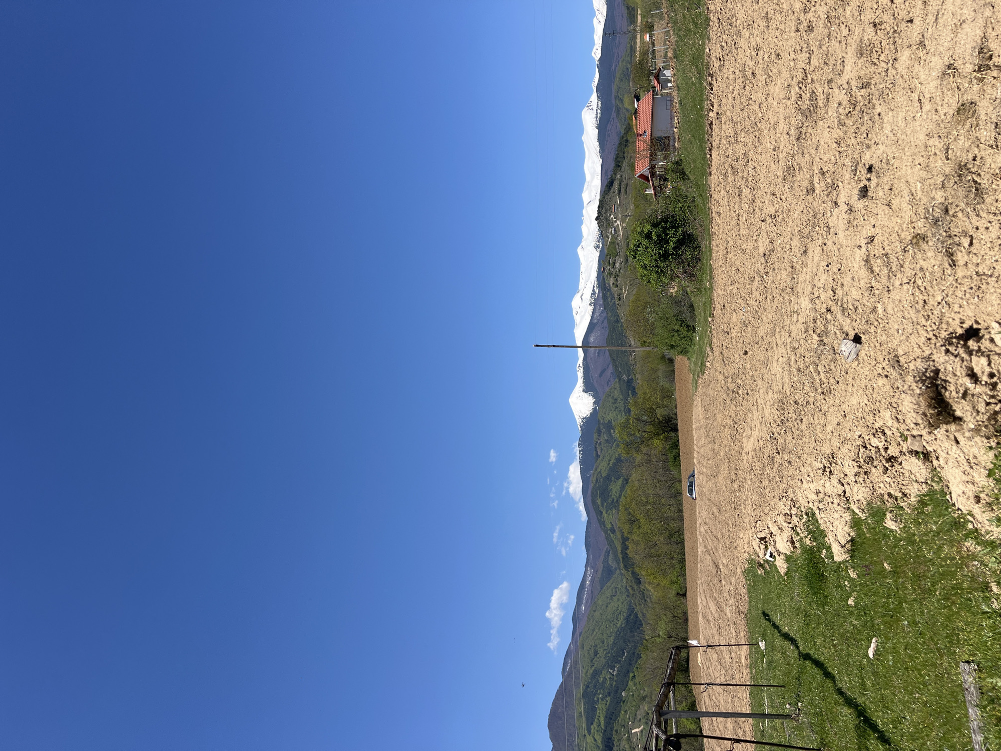 The image depicts a landscape in or near Kornitsa, Bulgaria. A clear, deep blue sky occupies the left half of the frame. On the right side of the image are grassy areas, dirt paths, and a cultivated field, suggestive of agriculture. In the mid-ground, vegetation and trees line the landscape near a ridge, leading towards snow-capped mountains in the distance. A structure with a red roof is visible amidst the vegetation. A utility pole is present, standing upright in the landscape. A fence is visible in the foreground.
