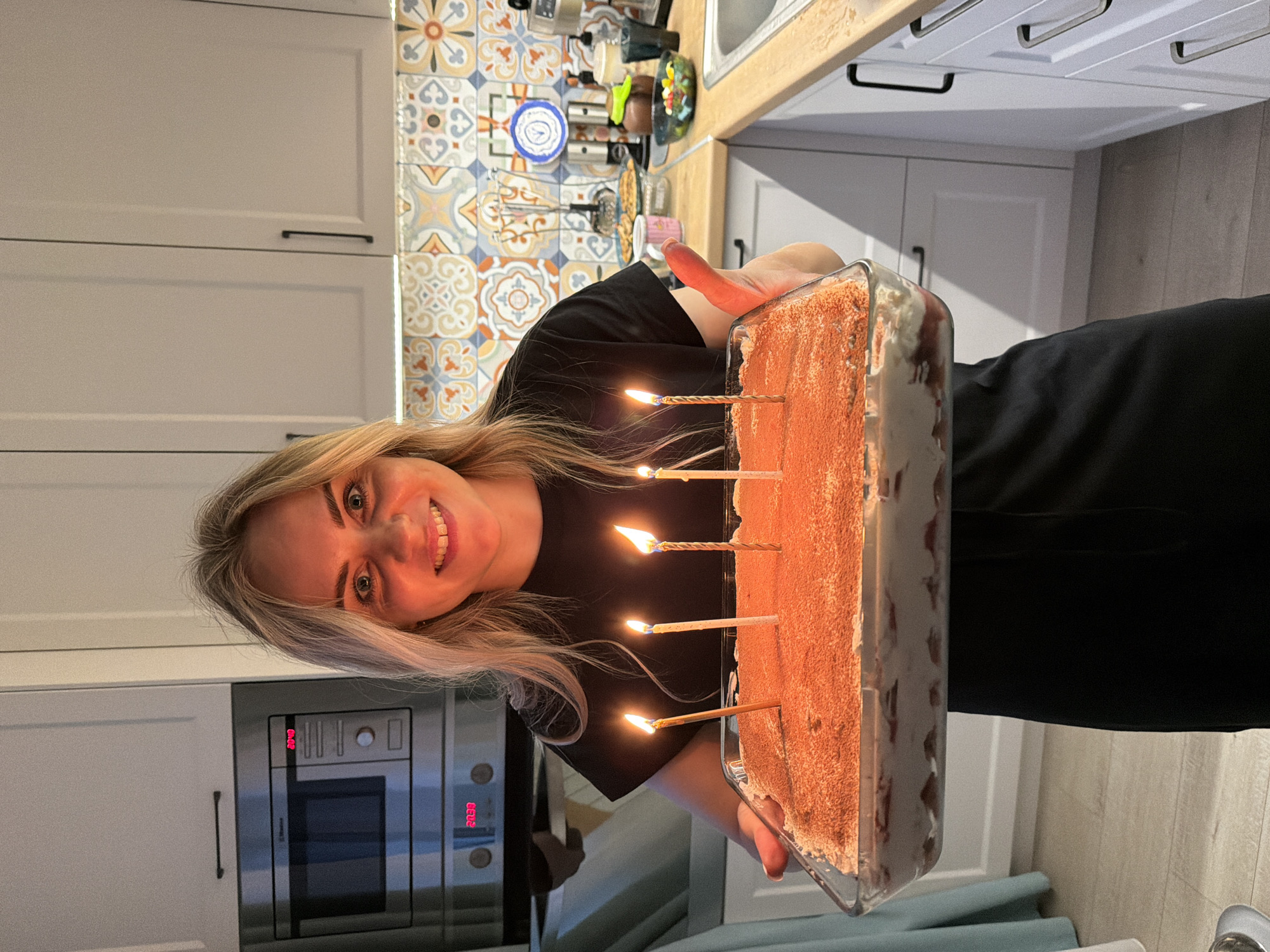 A fair-skinned woman with blond hair holds a rectangular glass baking dish filled with what appears to be a tiramisu cake, decorated with lit candles. The woman is smiling and appears to be indoors in a kitchen setting. The kitchen includes white cabinets with black handles, an oven with digital display showing "01.08", and a decorative backsplash with patterned tiles. Location: Kitchen interior, likely Votkinsk, Russia. No information is available to confirm the exact city.
