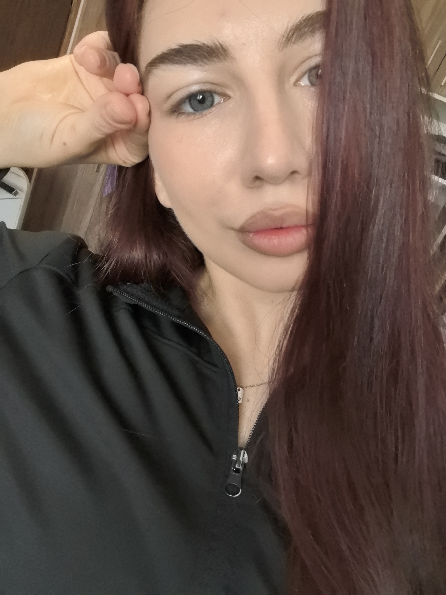A close-up, low-angle selfie shows a light-skinned woman with auburn hair that falls across the right side of her face. Her left hand is raised, with her fingers touching her forehead near her eyebrow. She has light blue eyes, prominent eyebrows, and full lips. She is wearing a black zip-up jacket with the zipper partially down, revealing a thin gold necklace with a small pendant. The background is out of focus, but appears to be an interior space with wooden elements. There is no discernible evidence in the image to definitively place the location as Stara Zagora, Bulgaria.