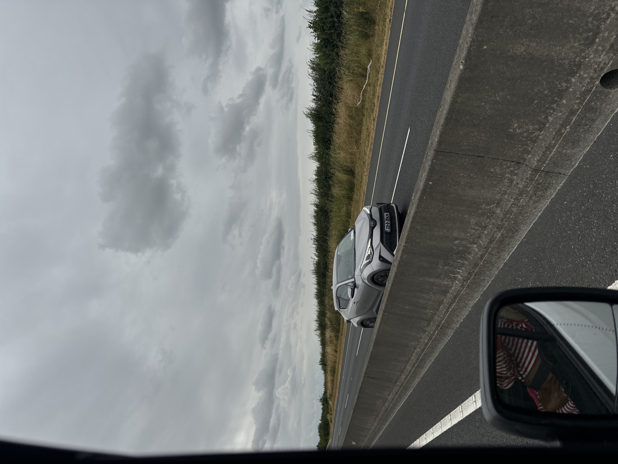 The image depicts a grey Toyota C-HR on a highway in Kilgowan Cottages, Ireland. The car is traveling on the right lane, with a concrete barrier separating it from the left lane. The sky is overcast with thick clouds. The surrounding environment features tall grass and bushes on the side of the road. A white plastic bag is visible snagged on the bushes near the road. The image is taken from a vehicle moving in the same direction as the Toyota.