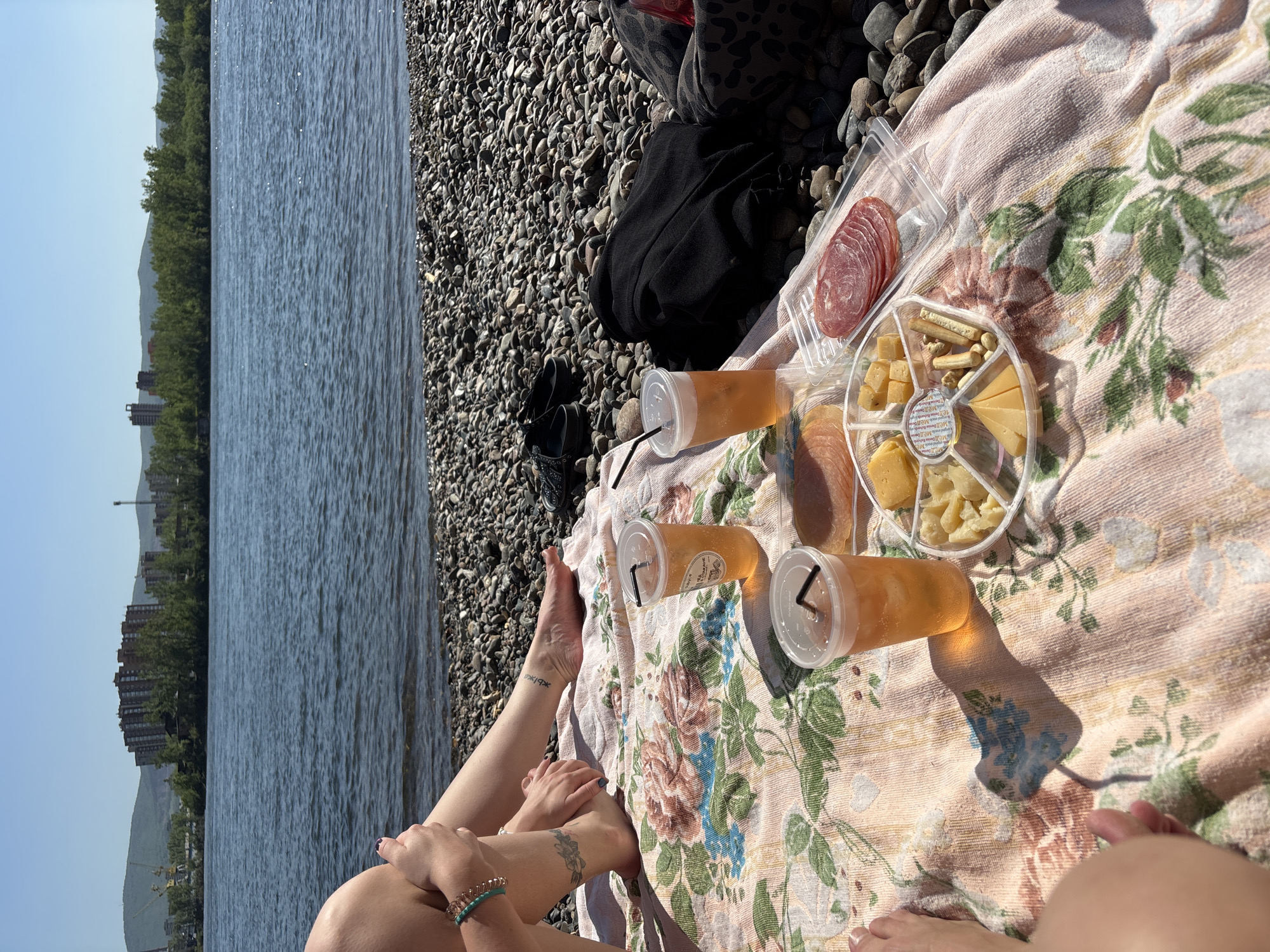 The image depicts a sunny outdoor picnic scene on a pebbled beach next to a large body of water, likely the Yenisei River in Krasnoyarsk, Russia. The foreground shows parts of at least two individuals, with their legs and feet resting on a pinkish floral-patterned blanket. One person's legs are prominently in the lower right, with painted toenails and a bracelet. Another person's legs are visible on the left, featuring tattoos, one appearing to be script (possibly Cyrillic or Hebrew) on the ankle and a floral design on the shin.
On the blanket, a selection of picnic items is laid out. These include three clear plastic cups with lids and black straws, containing an amber-colored beverage. Next to the drinks are snacks in clear plastic containers: one holds sliced salami or pepperoni, and another is a round, segmented platter filled with various cheeses (cubes and wedges), crackers, and nuts.
Beyond the beach, a wide expanse of calm, blue water stretches into the distance. The far shore is visible, lined with a dense treeline of green foliage, and behind it, several tall, multi-story buildings stand out against the landscape. Distant mountains or hills complete the background under a clear, bright blue sky.
The strong sunlight casts distinct shadows from the cups onto the blanket, indicating a clear, sunny day, likely during late afternoon. The scene conveys a relaxed and enjoyable atmosphere by the water. No distinct text is clearly readable on any items, though faint writing is present on a cup and a tattoo.