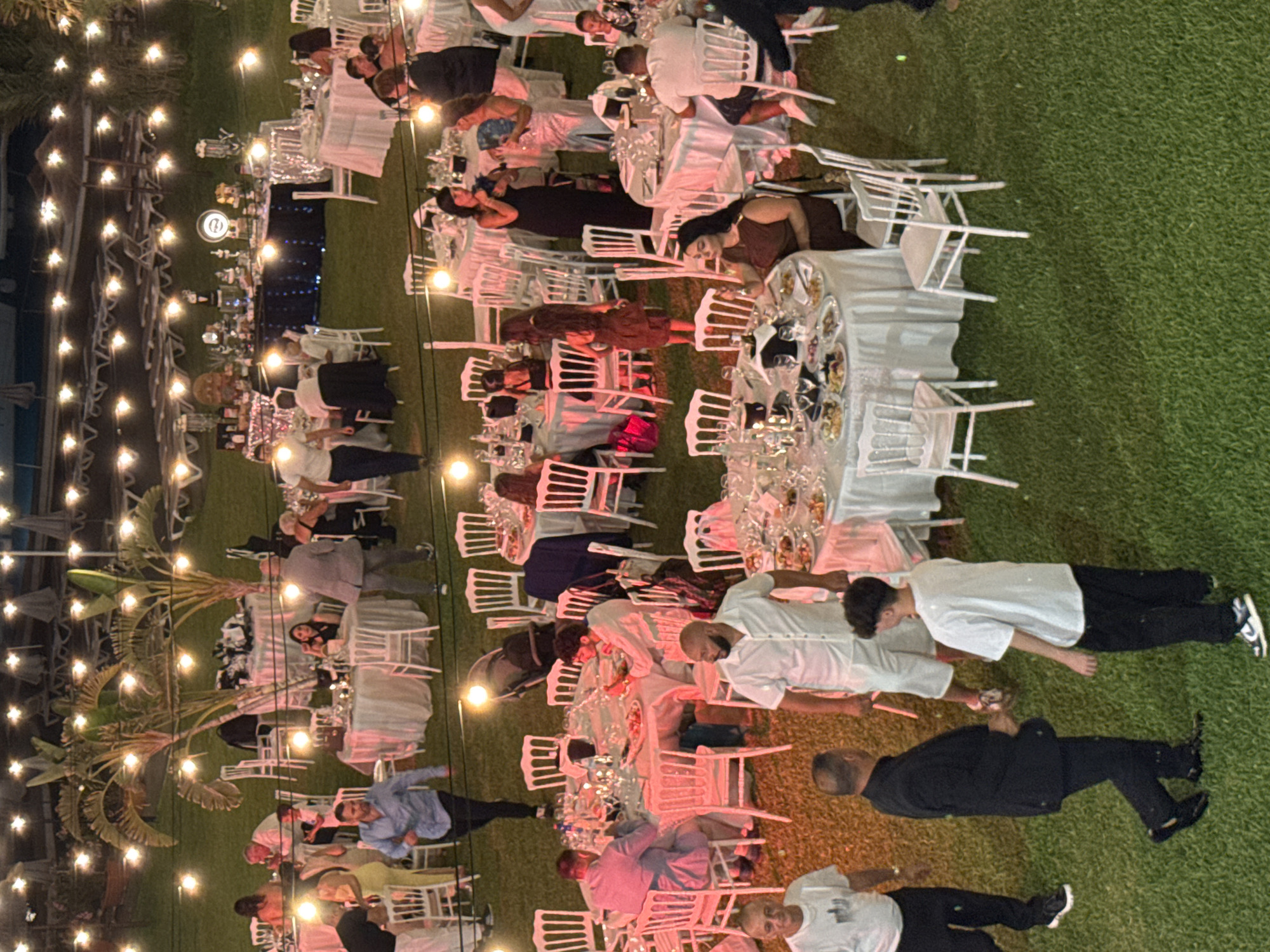 This is a high-angle, medium shot of an outdoor evening event held on a grassy area with many tables and chairs set up for dining. The scene is illuminated by numerous string lights suspended overhead, casting a warm glow. Palm trees with festive lights are visible, as are lounge chairs and umbrellas in the background, suggesting a resort or beachside location.
Several groups of people are present, dressed in semi-formal attire, engaged in dining and socializing around round tables covered with white tablecloths. The tables are laden with plates of food, glasses, and bottles. Some individuals are standing and interacting, while others are seated.
In the foreground, on the left, a man in a black suit and black pants is walking, talking to a young man in a white t-shirt and black pants. To the right, a man in a white short-sleeved shirt and white shorts is facing away from the camera, with other guests seated at tables behind him. Other individuals are visible throughout the scene, some in focus and others slightly blurred due to the lighting and distance. The overall impression is of a lively celebration or gathering.
Given the presence of palm trees and the outdoor setting with lights and lounge chairs, this scene is consistent with a venue in the city of Isola delle Femmine, Italy, known for its coastal resorts and evening events.