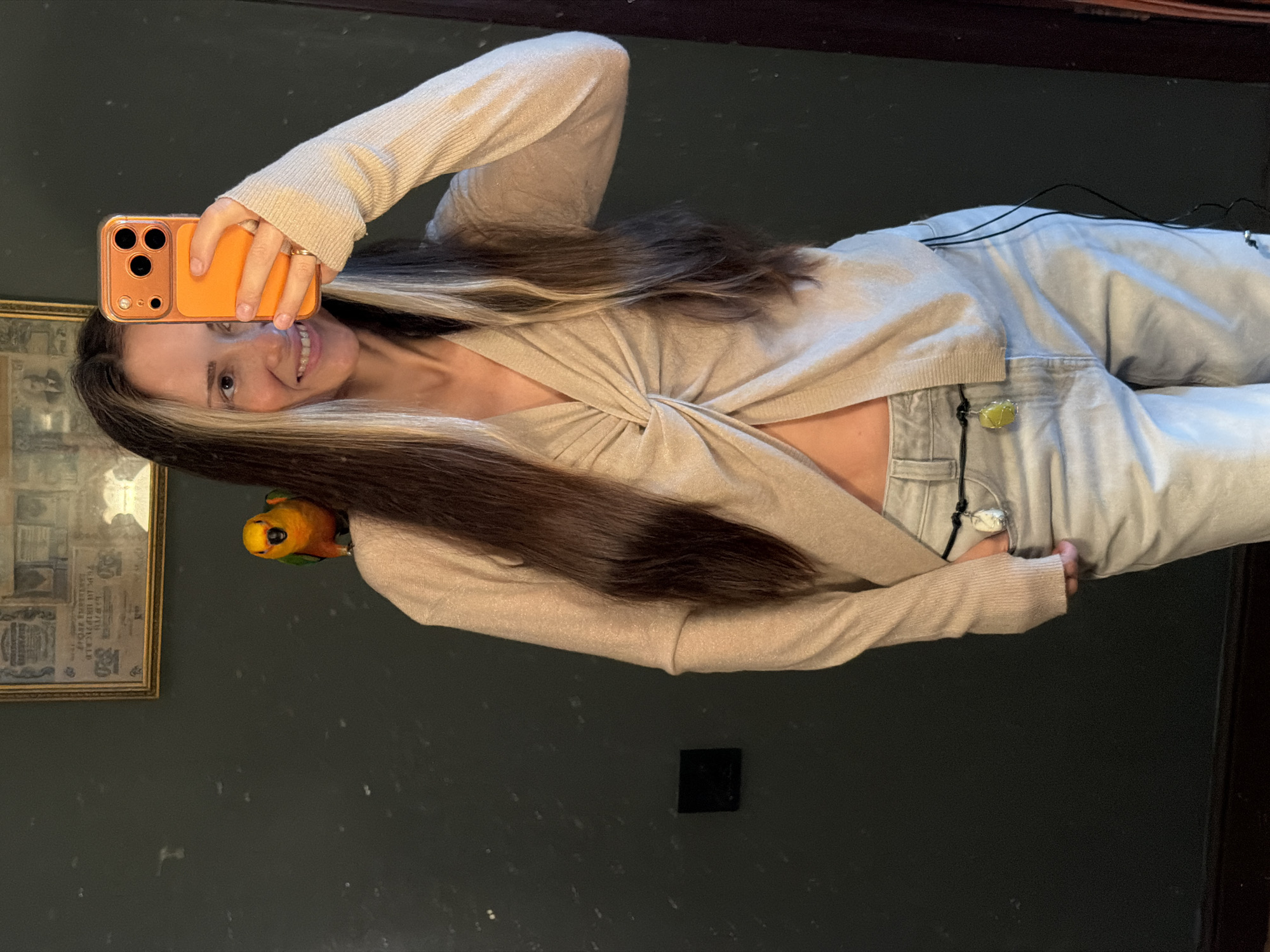 A woman with long brown hair, featuring blonde highlights, stands in front of a dark green wall. She is holding an orange smartphone in her right hand, taking a selfie. She is wearing a light beige, long-sleeved, knotted top that exposes her midriff, and light-wash jeans. A small, colorful parrot with orange and green feathers is perched on her left shoulder. The background includes a framed artwork on the wall and a mirror reflecting the scene. There is no indication that this image was taken in Khimki, Russia.
