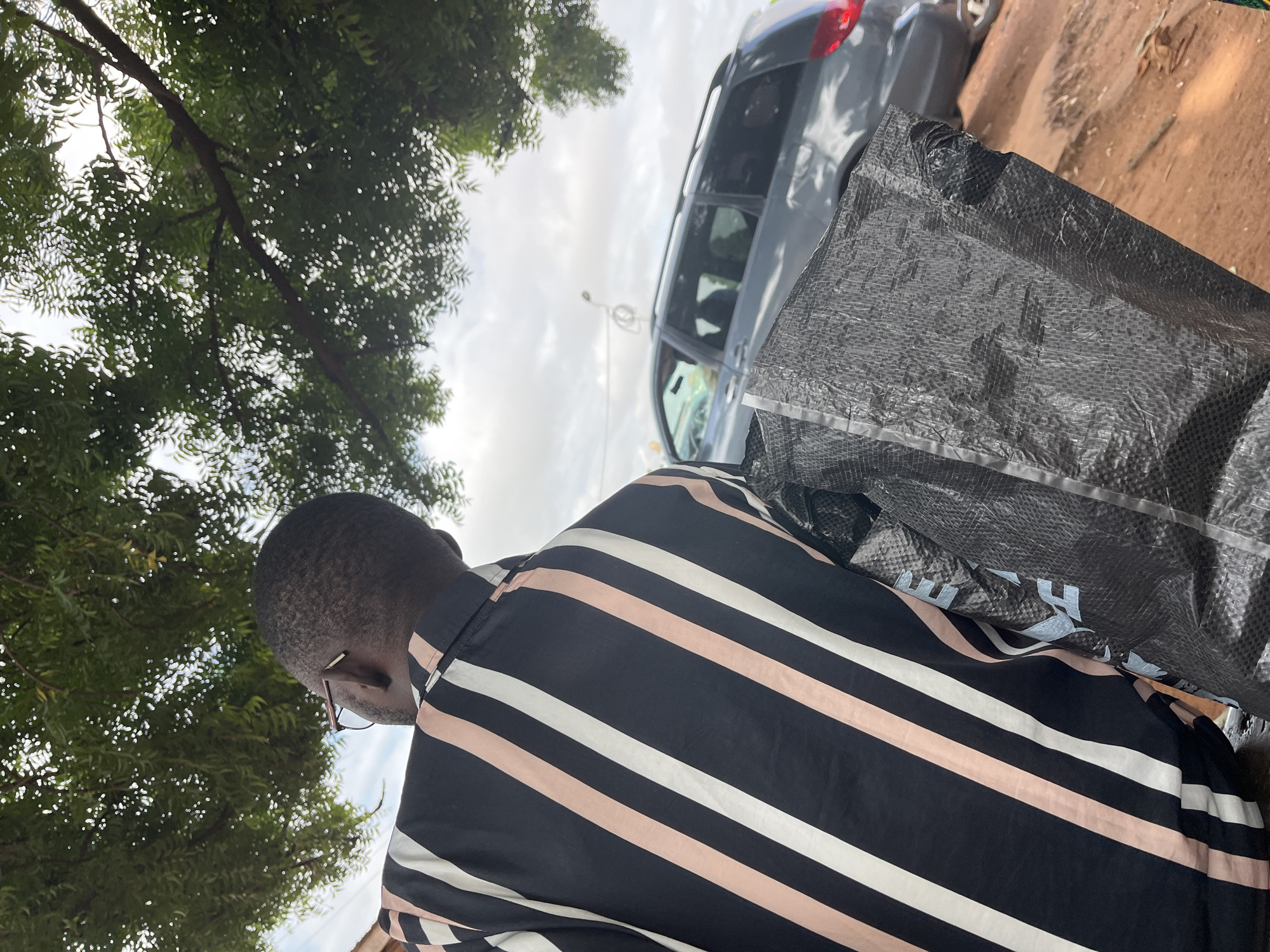 The image depicts an outdoor scene in Idah, Nigeria, viewed from slightly behind and below a person. A person with short, dark hair, wearing brown-framed eyeglasses, is seen from the back, facing towards the right side of the frame. They are wearing a black shirt with distinct vertical stripes in white and light peach/pink colors. The person is carrying a large, black, textured woven plastic bag, possibly a polypropylene sack, which occupies a significant portion of the lower right foreground. Light blue markings are faintly visible on the bag's surface.
In the mid-ground, a dark gray minivan or SUV with roof rails is partially visible, positioned on a reddish-brown dirt or soil surface that suggests an unpaved area or roadside. The vehicle's rear section, including a portion of its red taillight, is visible on the right.
The upper portion of the image is dominated by dense, green foliage from a large tree, with patches of bright, overcast sky visible through the leaves. The sky is predominantly cloudy with diffused light. A slender, light-colored pole or structure is faintly discernible in the distance beyond the vehicle. The overall lighting suggests natural daylight under a cloudy condition.