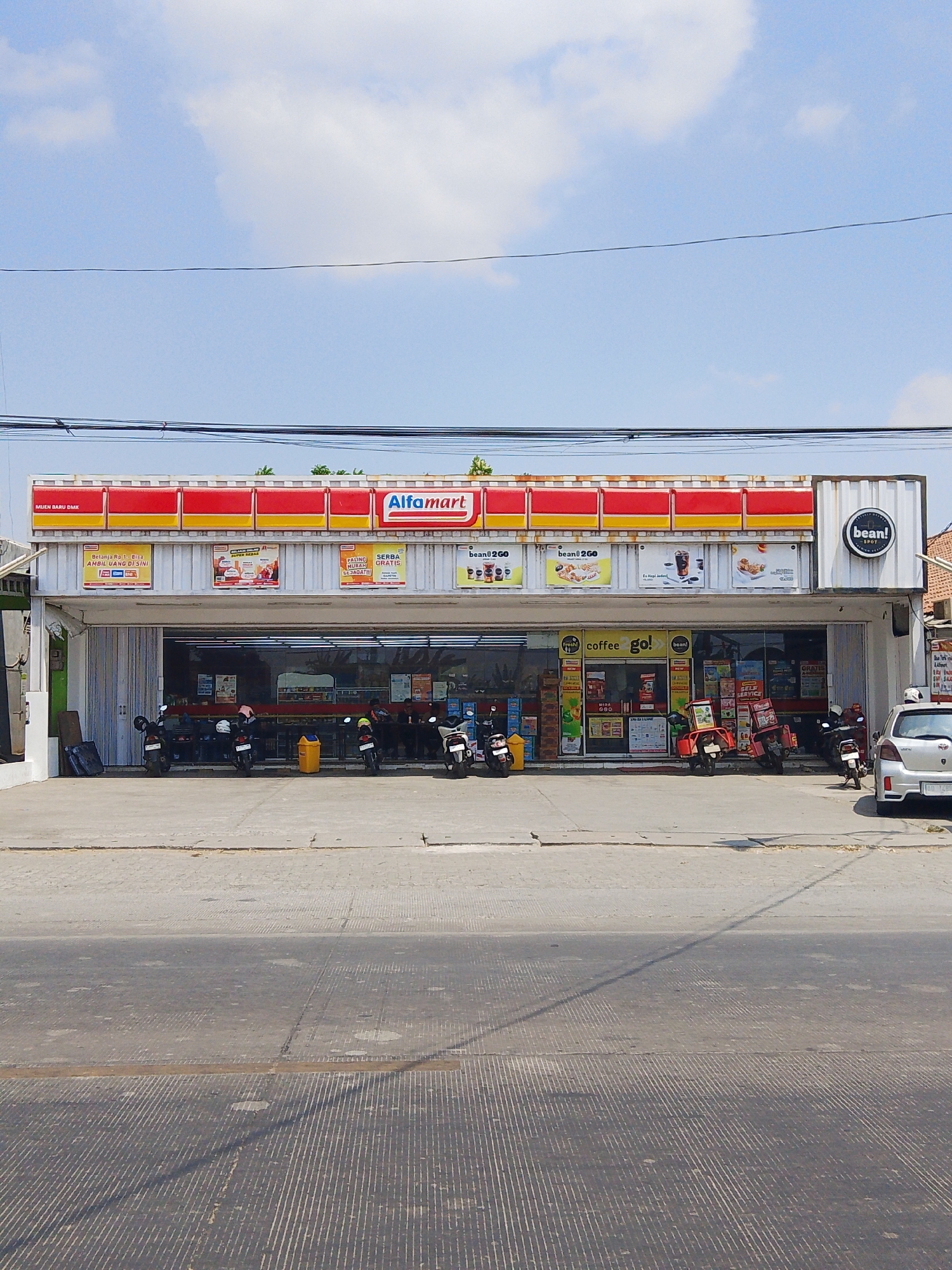A daylight, eye-level, medium shot shows the facade of an Alfamart convenience store in Bengkal, Indonesia. The store has a red and yellow striped awning and large glass windows. Various promotional signs are visible on the exterior walls. Several motorcycles are parked in front of the store, and a silver car is parked to the right. The street in front of the store is paved with concrete and has a striped pattern. The sky is blue with some clouds.