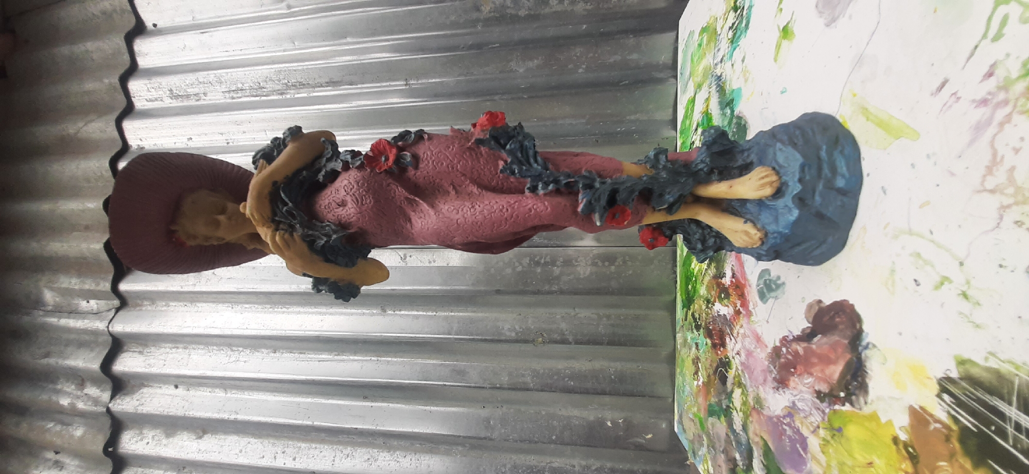 The image shows a statue of a woman with a large hat and a flowing pink dress adorned with vines and red flowers. The statue stands on a blue base and is placed on a surface splattered with various colors of paint. Behind the statue is a corrugated metal wall. The scene appears to be indoors, possibly in a workshop or studio, with the paint splatters suggesting an artistic or crafting environment. The statue is centrally positioned and in focus, while the background elements are less distinct. There is no discernible information about the city of Yessentukskaya, Russia, within the image itself.