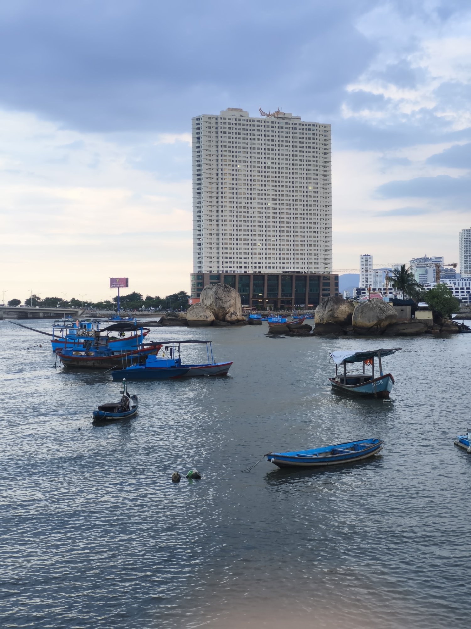 This image depicts a harbor scene in Bac Nha Trang Ward, Vietnam. A tall, modern white high-rise building dominates the background, its facade covered in numerous windows. In the middle ground, several blue fishing boats of various sizes are moored in the calm water. Some of the boats have canopies, and one in the foreground has a white tarp. A rocky outcrop with a small structure and a palm tree is visible to the right of the boats, with more buildings and cranes in the further distance. The sky is overcast with soft, diffused light, suggesting it is either early morning or late afternoon. The water is a deep blue-gray, reflecting the cloudy sky. There are no prominent activities taking place, with the boats appearing to be at rest. A few buoys are visible in the water, along with a person who appears to be swimming or floating. On one of the boats, a license plate with the inscription "VN-174-TD" is partially visible. In the distance, a bridge spans the water, with trees and streetlights lining the shore. The overall mood is peaceful and serene.
