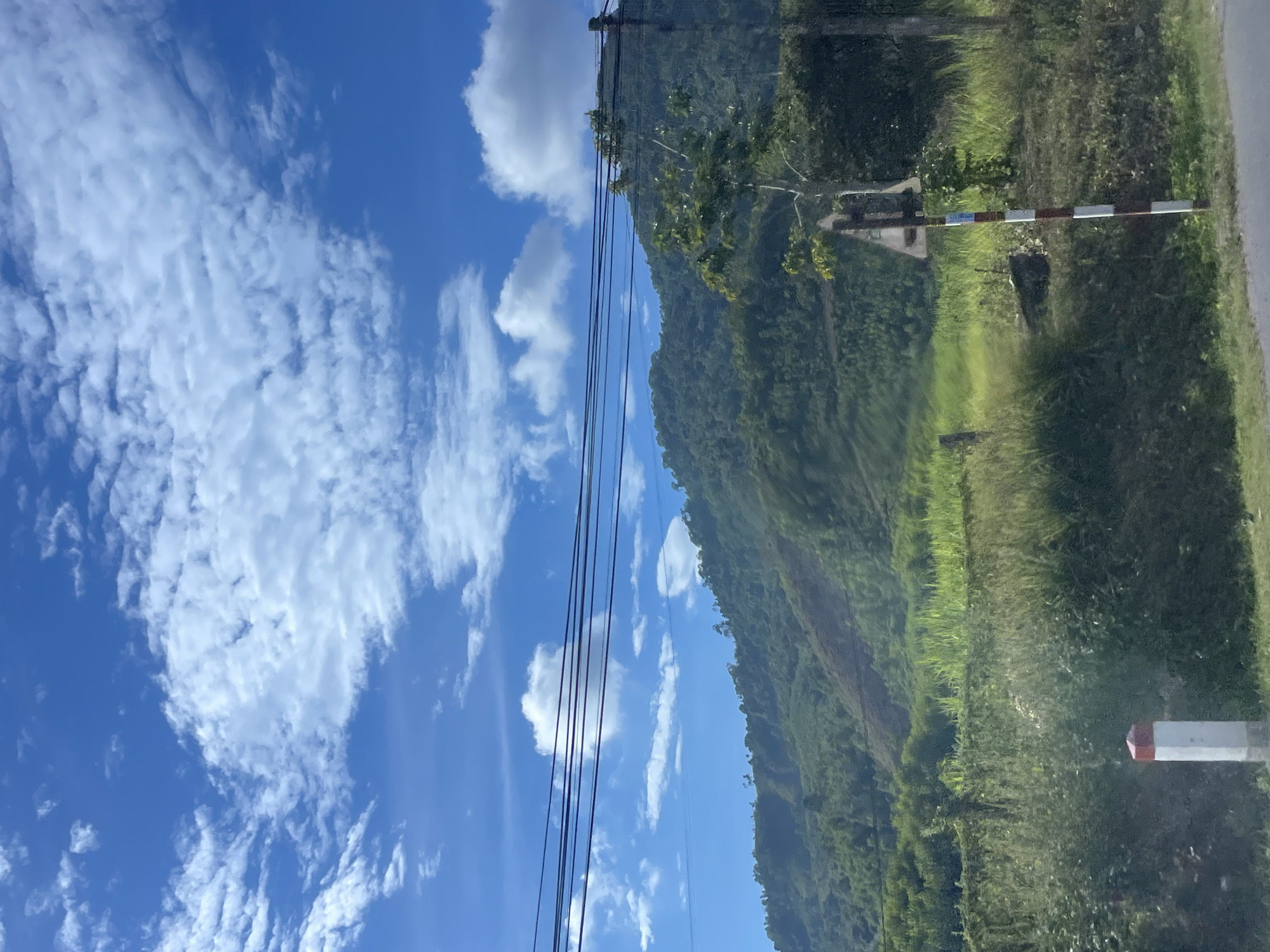 The image depicts a view of a lush, green mountainside under a bright blue sky with scattered white clouds. Power lines are visible strung across the upper portion of the frame, running horizontally. In the foreground, to the right, is a white and red striped pole with a triangular sign attached to it, possibly a road marker. Another similar white and red marker pole is partially visible on the left side of the frame. The landscape appears rural, with dense vegetation covering the hills and grassy areas in the foreground. The scene suggests a bright, possibly sunny day in a natural, mountainous setting. The context provided indicates this location is in Hướng Hiệp Commune, Vietnam.