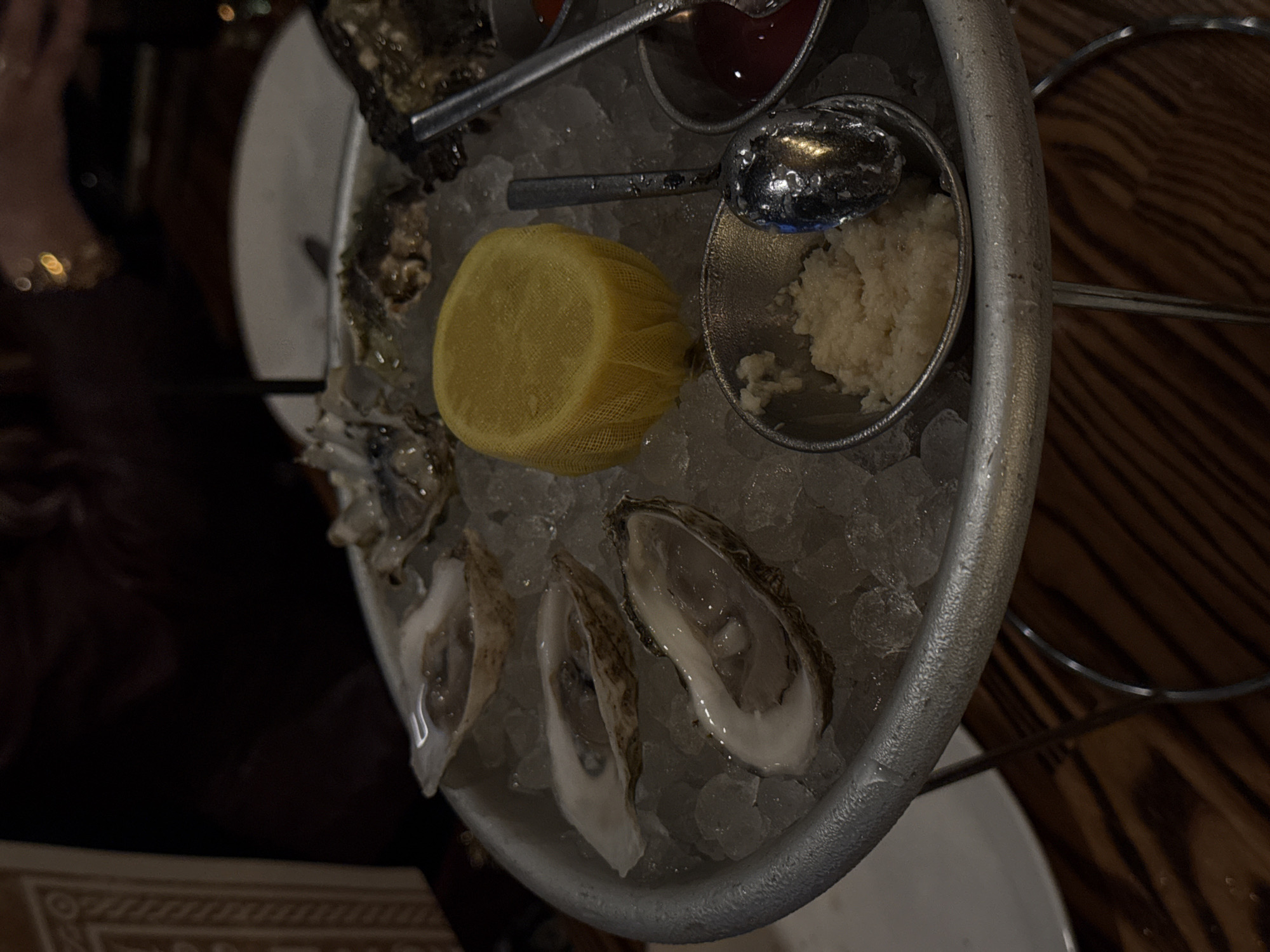 A slightly elevated, top-down, angled shot depicts an ice-filled, round, metal oyster platter with oysters, a lemon wedge in netting, horseradish, and cocktail sauce, situated in a dimly lit restaurant.
The silver platter contains several shucked oysters arranged on crushed ice. At the top of the platter, directly above the oysters, are two small metal containers, one filled with a dark red sauce and the other with a white, grated substance. A metal spoon rests on top of the white substance. A lemon wedge encased in a yellow netting material sits near the center.
The table to the left of the platter has a partially visible person's arm and hand with a ring. The table underneath the platter is partially visible in the bottom half of the image. The background features a mix of dark, out-of-focus forms and a horizontally patterned wood surface on the right.
Based on common knowledge, the image appears to be taken in a restaurant, potentially in San Diego, USA. Specific details such as oyster type or exact location within San Diego are not discernible.