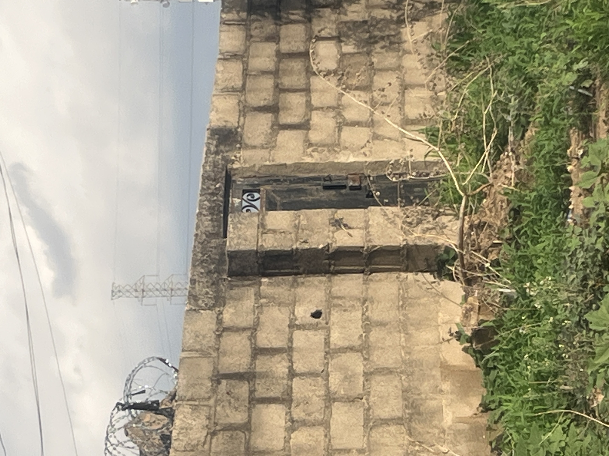 The image shows a partial view of a masonry structure in Barakin Akawo, Nigeria. The structure is constructed from rectangular concrete blocks. There's an opening in the wall, partially covered by a dark metal grate or door with decorative wrought iron elements. An object appears to be present, or affixed within the wall interior, near the opening’s iron details. The edges of the structure are overgrown with vegetation. In the background, an electric power transmission tower and barbed wire are visible against a cloudy sky.