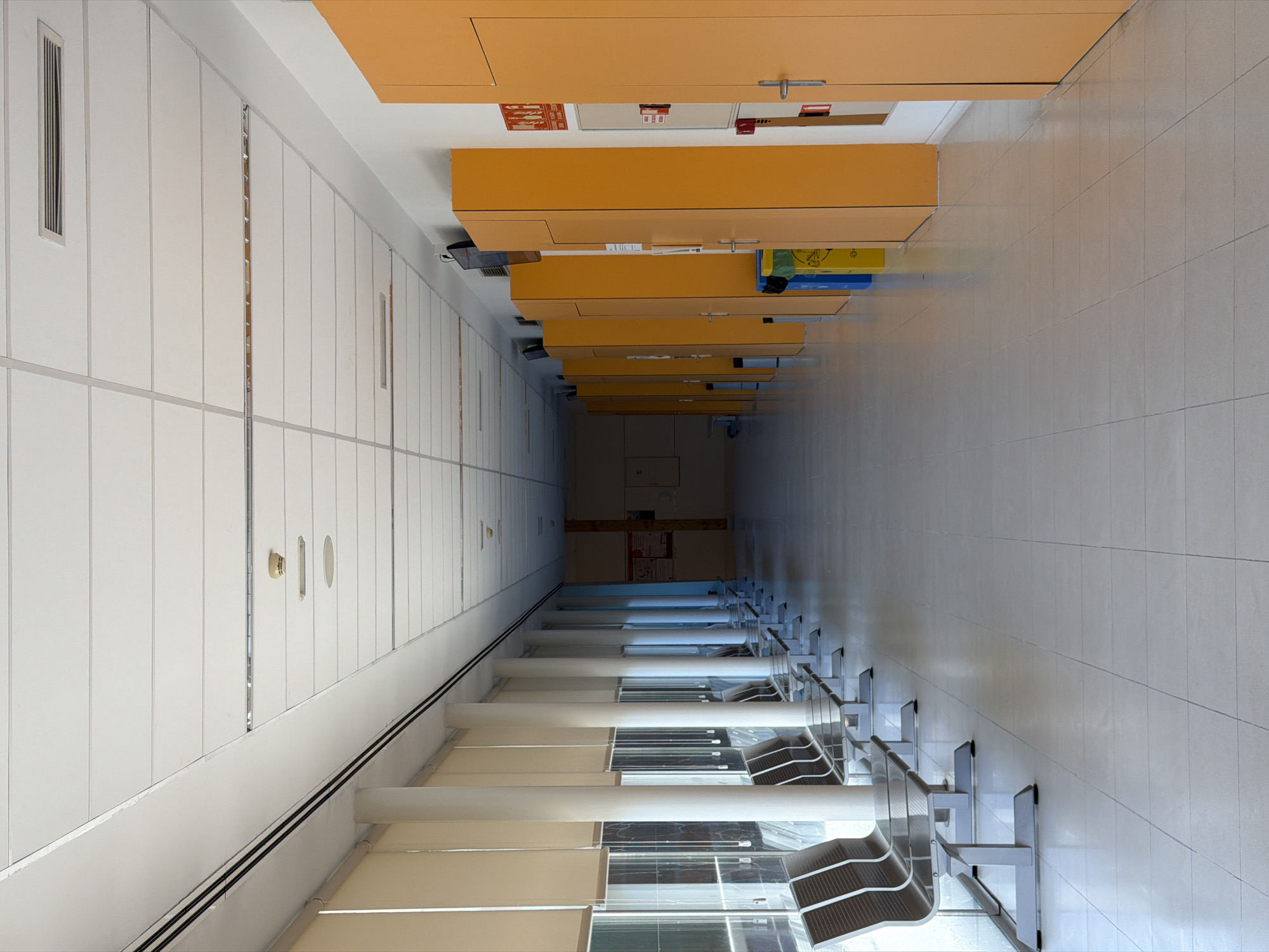 This is a forensic, matter-of-fact description of a hallway in a public building, possibly a clinic or office, located in Las Rozas de Madrid, Spain.
The hallway stretches into the distance, with a polished, light-colored tiled floor. To the left, a row of floor-to-ceiling windows is covered by beige roller blinds, with large white cylindrical columns spaced intermittently between them. A series of metallic benches with dark seating are positioned along the wall near the windows, with glass tabletops attached to the benches.
To the right, a long wall is lined with tall, bright orange cabinets or doors, punctuated by silver handles and what appear to be key card readers or intercoms. Beside the cabinets, there are two trash receptacles, one blue and one yellow. A fire extinguisher sign and a fire alarm are visible on the wall near the foreground right.
The ceiling is a modular grid of white acoustic tiles with recessed linear vents and a circular light fixture. A smoke detector is also visible on the ceiling. The lighting is bright and even, suggesting artificial illumination. The hallway appears clean and orderly, with no people present. The overall impression is one of sterile functionality.