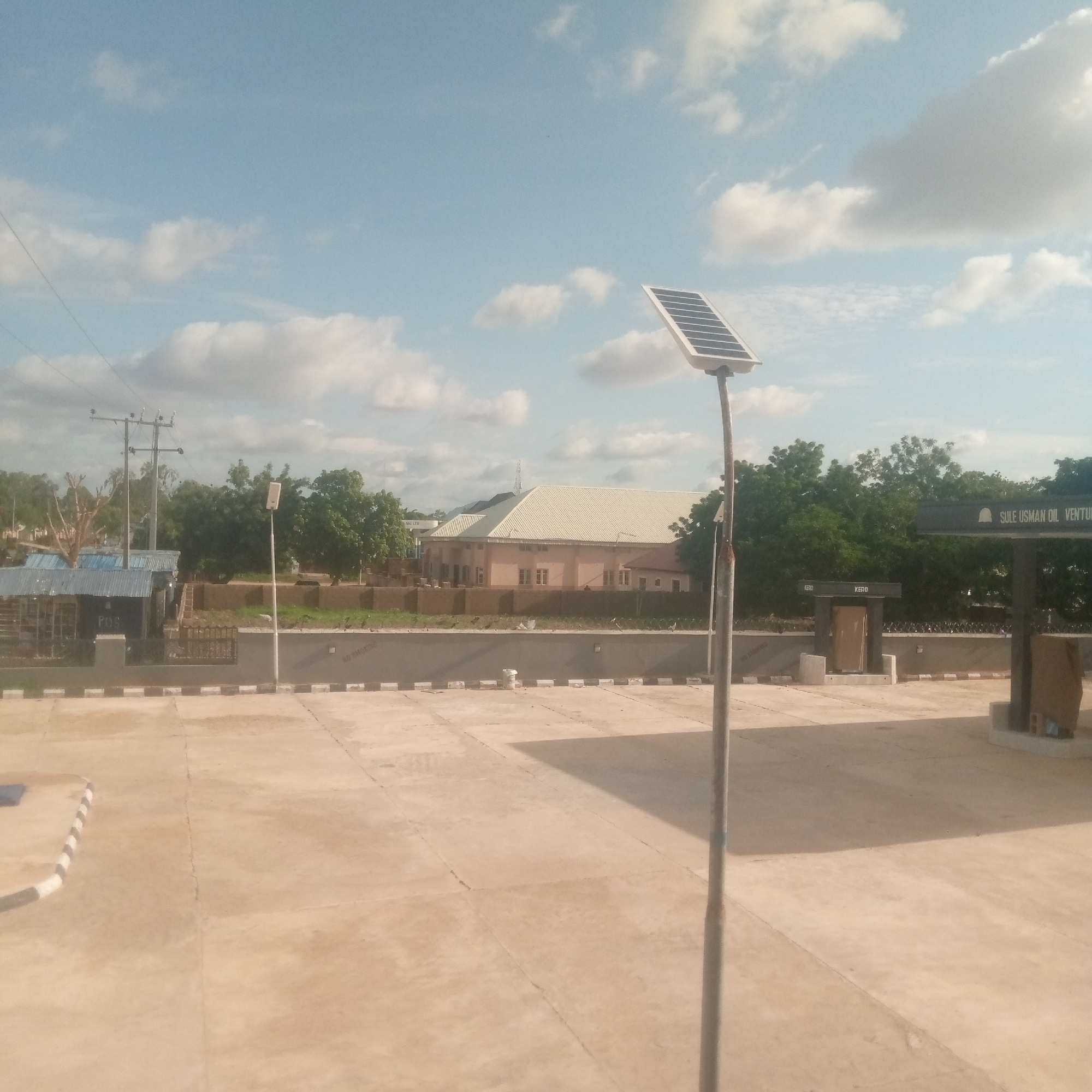 The image depicts an outdoor scene under a bright, partly cloudy sky during daylight hours. The foreground is dominated by a large, light tan-colored concrete paved area with visible expansion joints and minor staining. Black and white striped curbing is visible along the lower left edge and bordering a dark gray wall that extends across the mid-ground.
In the right mid-ground, a small solar panel is mounted on a slender pole that shows signs of rust or weathering, angled upwards towards the left. To the immediate right, a dark canopy structure bears the white text "SULE USMAN OIL VENTURE," indicating a fuel station. Below this, two dark kiosks or enclosures are visible, each with the word "KERO" in white above their entrances, suggesting a kerosene dispensing point.
The dark gray wall running across the mid-ground is topped with a black metal fence and has the white stenciled phrase "NO SMOKING" repeated at intervals. Behind this wall, in the central background, a prominent two-story building with light peach-colored walls and a large, pitched white corrugated metal roof stands among green trees.
To the left mid-ground, a white-painted pole supports an LED-style streetlight. Further left, utility poles with multiple cross-arms and power lines are visible above a smaller, low building with a blue corrugated metal roof. The faint text "POS" is discernible on the front of this smaller building. A bare, deciduous tree stands between this building and the central background building. Dense green foliage and numerous trees are scattered throughout the background, obscuring the horizon.
No individuals or vehicles are visible within the frame. The scene appears to be a commercial property, specifically a fuel station, in a potentially developing or semi-urban area. The exact city and state are unknown, but the naming convention suggests a location within Nigeria or a similar West African country.