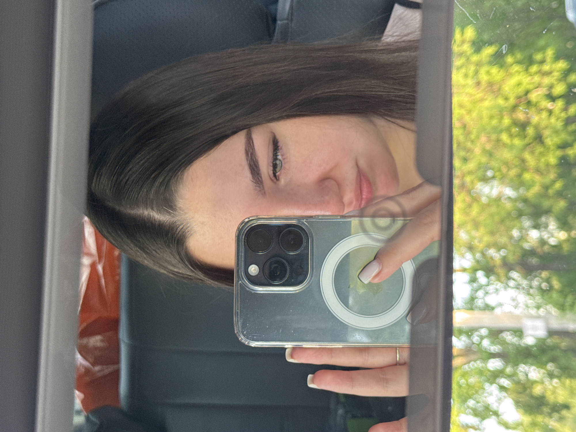 A person is taking a selfie in a car mirror. The individual has long brown hair and is holding a cell phone with a transparent case. In the reflection of the mirror, a blurred view of green trees can be seen outside the car window. The interior of the car is also visible, including the dark seats. The image doesn't provide enough information to determine the exact location in Perm, Russia.