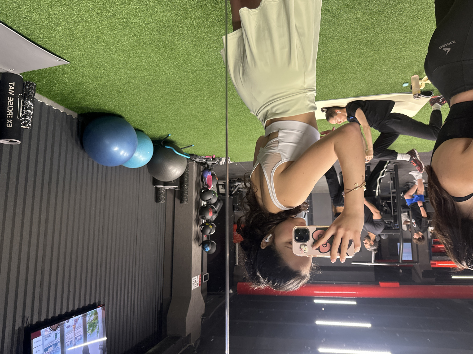 A selfie is taken in a mirror in what appears to be a gym setting. The person taking the selfie is wearing athletic wear, and is holding a phone with a decorated case. Several other individuals are present in the background, engaging in various exercises. Gym equipment, including free weights and exercise balls, can be seen. The setting appears to be indoors, with artificial lighting. The gym appears to be clean and well-maintained. Given the context of other gym users, the location is likely a fitness center. There is no specific information available to determine whether this is in Yerevan, Armenia, or to determine any specific locations.
