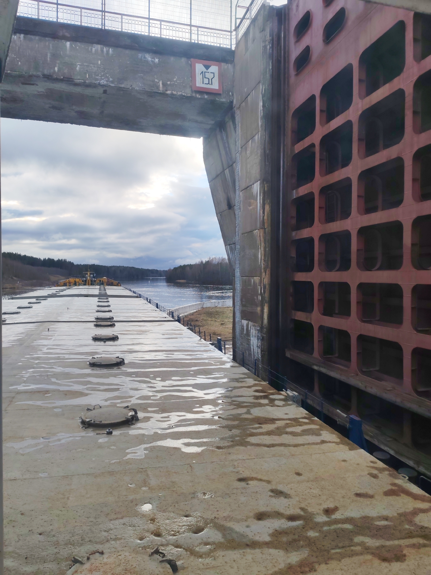 The image shows a large canal lock. The lock has a metal gate on the right with large square openings. The number "15.7" is visible on a sign attached to the concrete structure above the passageway. A wet concrete surface stretches out towards the horizon, punctuated by round metal hatches. In the distance, a barge is visible on the water, surrounded by trees. The sky is overcast.