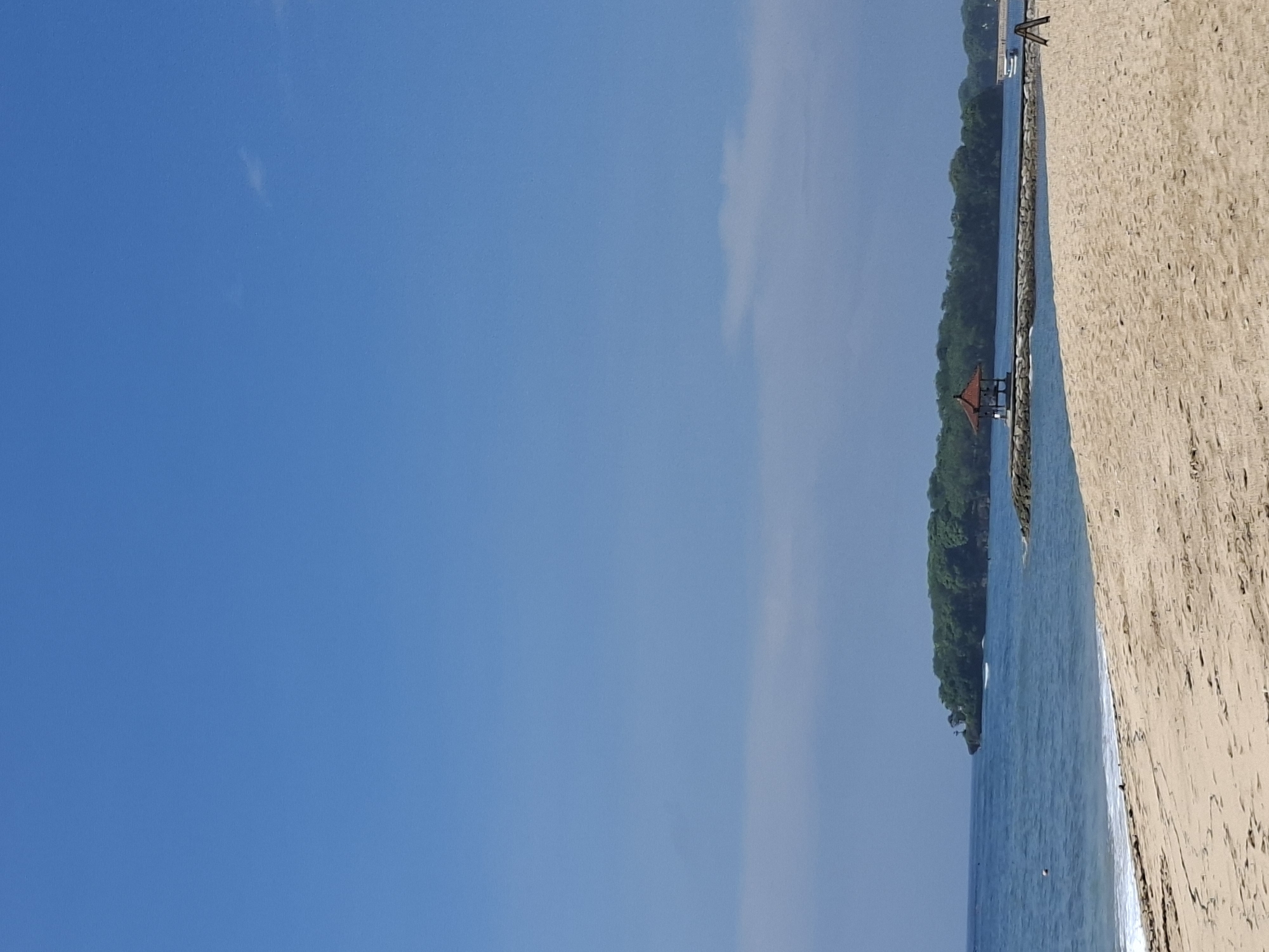The image shows a sunny beach scene in Kuta Selatan, Indonesia. The sky is clear blue with a few wispy clouds. A sandy beach occupies the right side of the frame. The ocean runs along the bottom center, separated from the beach by a shoreline. A narrow strip of land covered in lush green vegetation separates the ocean from what appears to be another body of water. There is a small red-roofed structure on the strip of land. A long stone structure is visible along the shoreline. At the far end, something that seems to be a ladder can be seen.