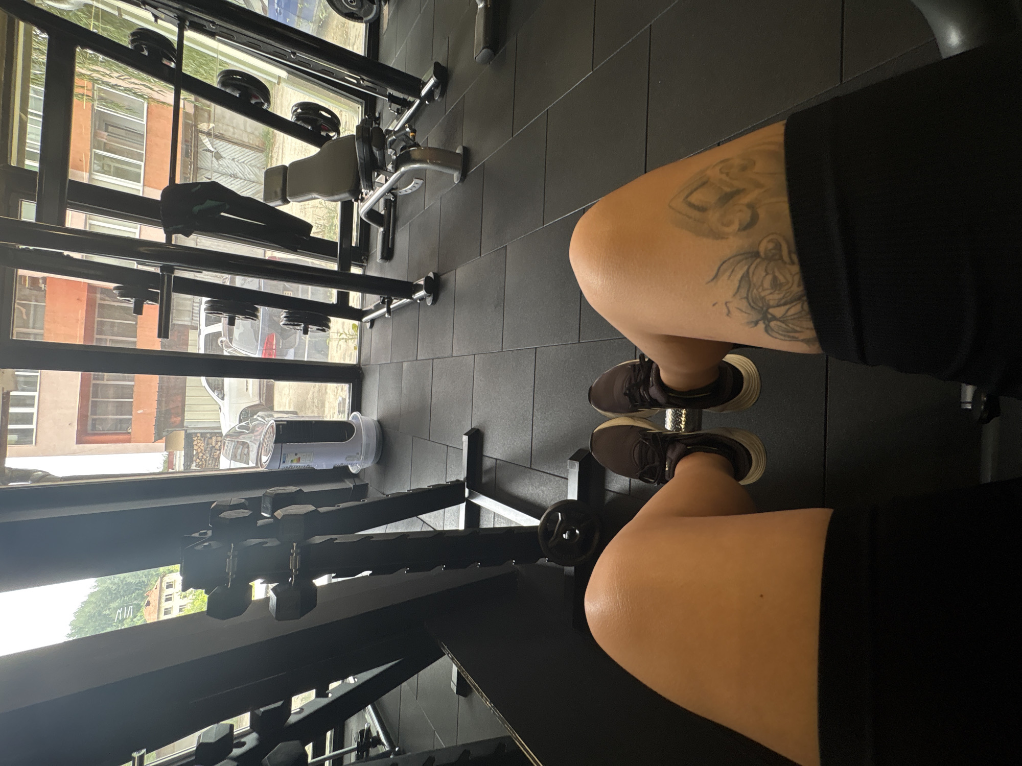 The image depicts a POV shot from a person's perspective within a gym in Troyan, Bulgaria. The foreground is dominated by the individual's legs, clad in black shorts and athletic shoes, resting on what appears to be a weight machine. The person's left thigh bears a tattoo of a flower and geometric shapes. In the midground, various gym equipment is visible, including weight racks, dumbbells, and a bench press. The background reveals a large window looking out onto a street scene in Troyan, with buildings, trees, and parked cars visible. The gym flooring consists of black rubber tiles.