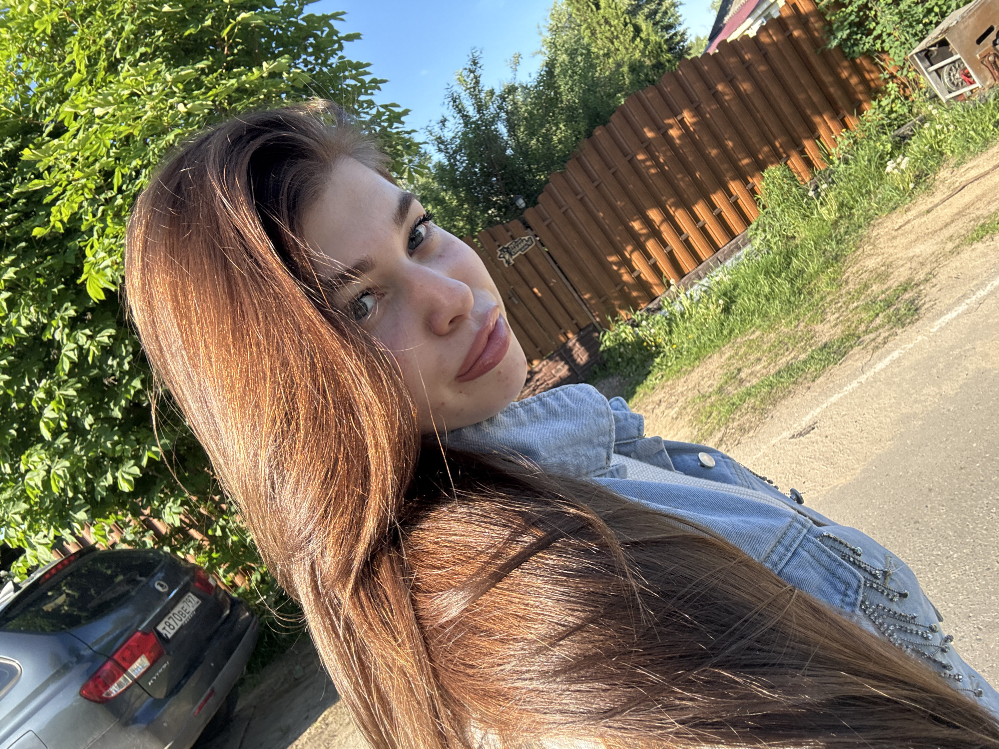 A young woman with long, brown hair is facing the camera with a slight tilt of her head. She is wearing a light blue denim jacket. Behind her are green trees and a brown fence. A gray car with a license plate is visible to the left of the woman. The setting appears to be a road or sidewalk in a suburban or rural area. The location is in Мещёры, Russia.