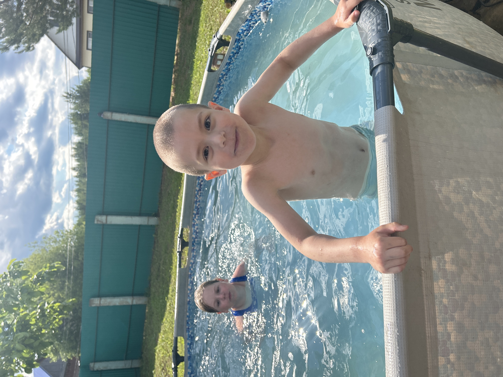 In an outdoor setting, two children are enjoying a swim in an above-ground pool. The pool has a patterned liner visible along its edge. The water is clear and blue, with sunlight reflecting off its surface, indicating a sunny day.
The foreground is dominated by a young boy, who appears to be leaning into the frame and looking directly at the camera with a cheerful expression. He is shirtless, and his hand is gripping the edge of the pool. Behind him, another child, a girl, is visible swimming in the pool. She is wearing a blue swimsuit.
The background shows a green metal fence that encircles the property, with grass and trees visible beyond it. A house with light-colored walls and windows can be seen over the fence. The sky is partly cloudy with bright sunlight. The overall atmosphere is one of summer fun and outdoor recreation. The location context provided is Foky, Russia. There is no visible text in the image.