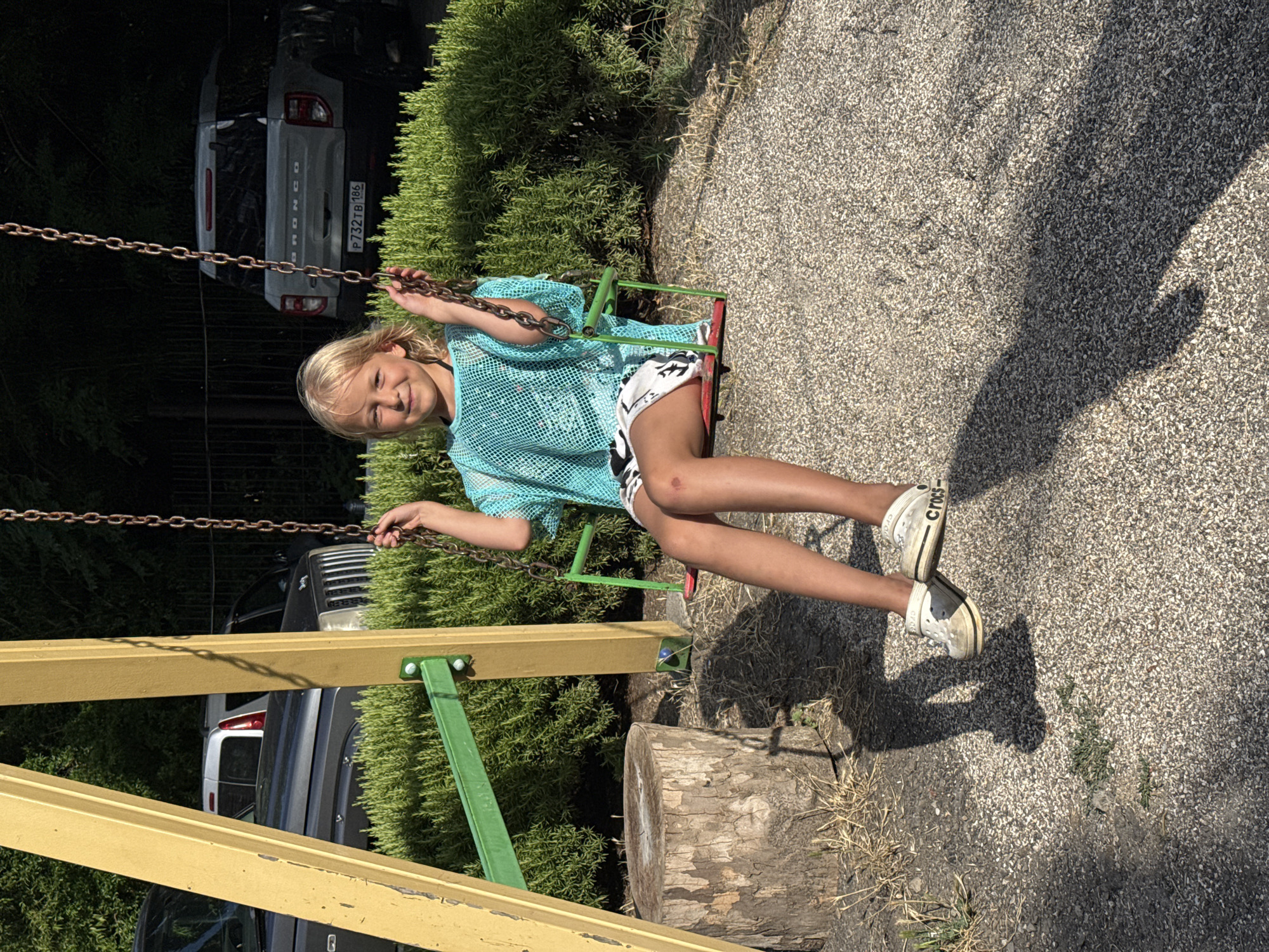 A young blonde girl is sitting on a swing, smiling at the camera. She is wearing a light blue mesh top and white shorts with a black pattern. Her shoes are white Crocs with black trim. Her right knee has a scrape on it. The swing set is yellow and green, and it's located in a playground with a gravel surface. Behind the swing set, there is a dense green hedge. In the background, several cars are parked, including a silver Ford Bronco with a Ukrainian license plate. The scene appears to be in a park or a residential area. The time of day is sunny, with shadows cast on the ground.