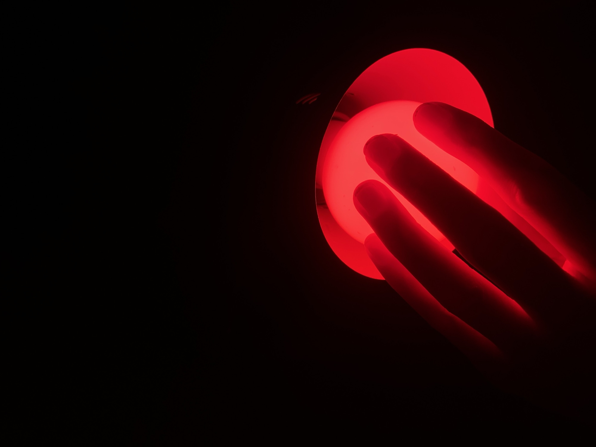 A hand with three fingers illuminated by a bright red light is positioned over a glowing red circular object against a black background. The fingers are positioned to the left of the circle, with the fingertips touching the edge of the illuminated surface. The overall scene suggests a close-up view of interaction with a light source. There is no discernible information about the city of Budva, Montenegro, within the image content.
