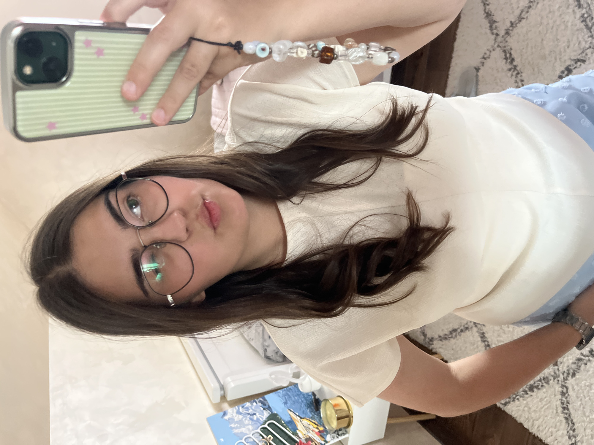 This is a medium shot, selfie-style photograph of a young woman with dark brown hair and glasses, taken indoors. She is wearing a light-colored, short-sleeved top and blue patterned bottoms. In her right hand, she is holding a smartphone with a green and white striped case decorated with pink stars, and a beaded charm is attached to the phone. The woman is looking at the camera with her lips pursed. To her left, a nightstand is visible with a gold cylindrical object and a framed picture depicting a landscape. The background shows a bed with white bedding and a patterned rug on the floor. The lighting appears to be natural, coming from an unseen window. There is no indication of the city of Koroviia, Ukraine.