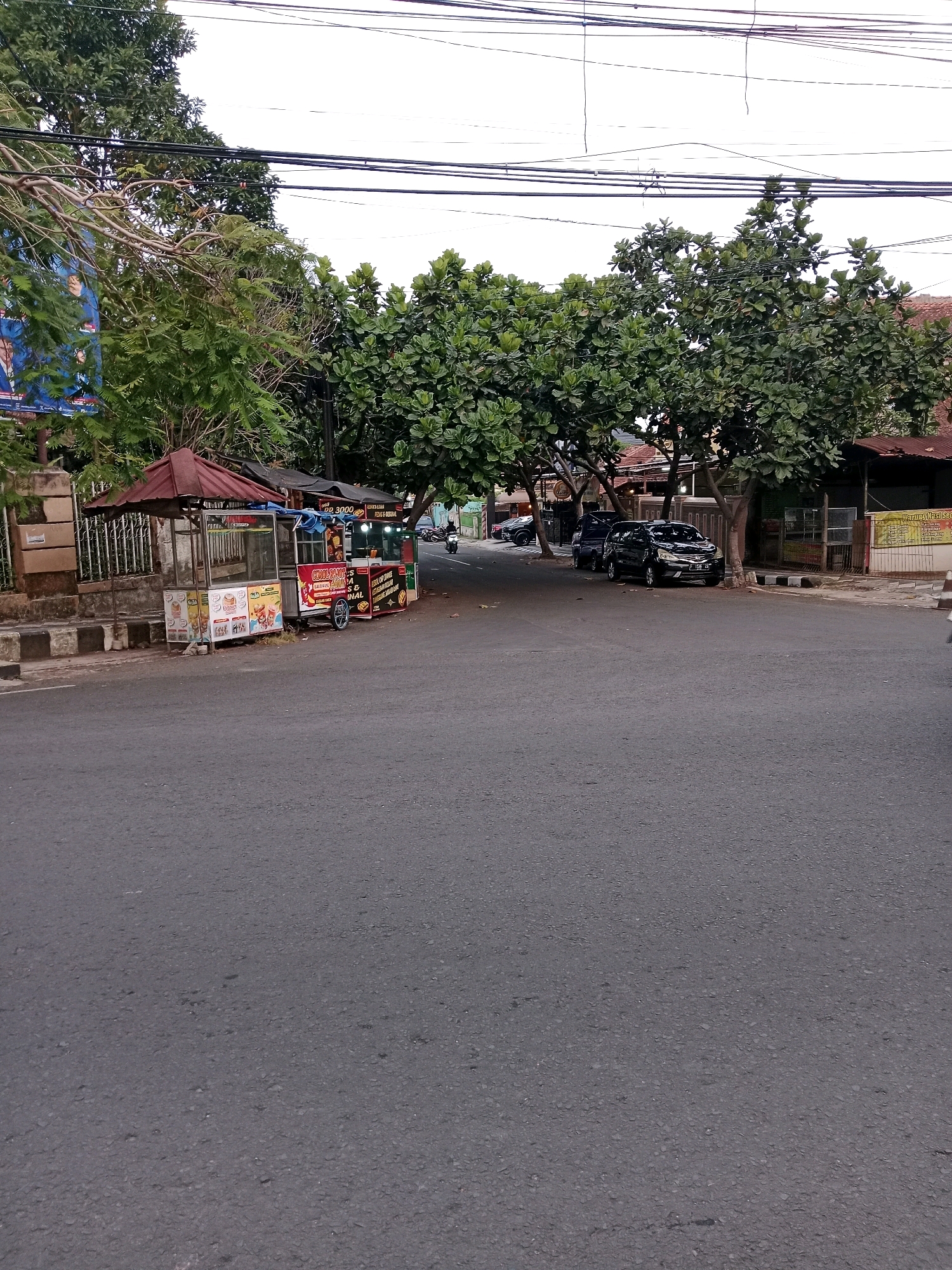 The image depicts a street scene in Cianjur, Indonesia. In the foreground is an asphalt road. Along the side of the road, there are two food carts, each with a maroon awning. The carts are displaying food items and pricing, with text in Indonesian. Behind the food carts, there are trees with full green foliage. Several black cars are parked along the side of the road under the trees. In the distance, a motorcycle is visible on the road. Overhead, electrical wires are strung across the sky. The environment appears to be a typical street in a populated area, with buildings visible in the background.