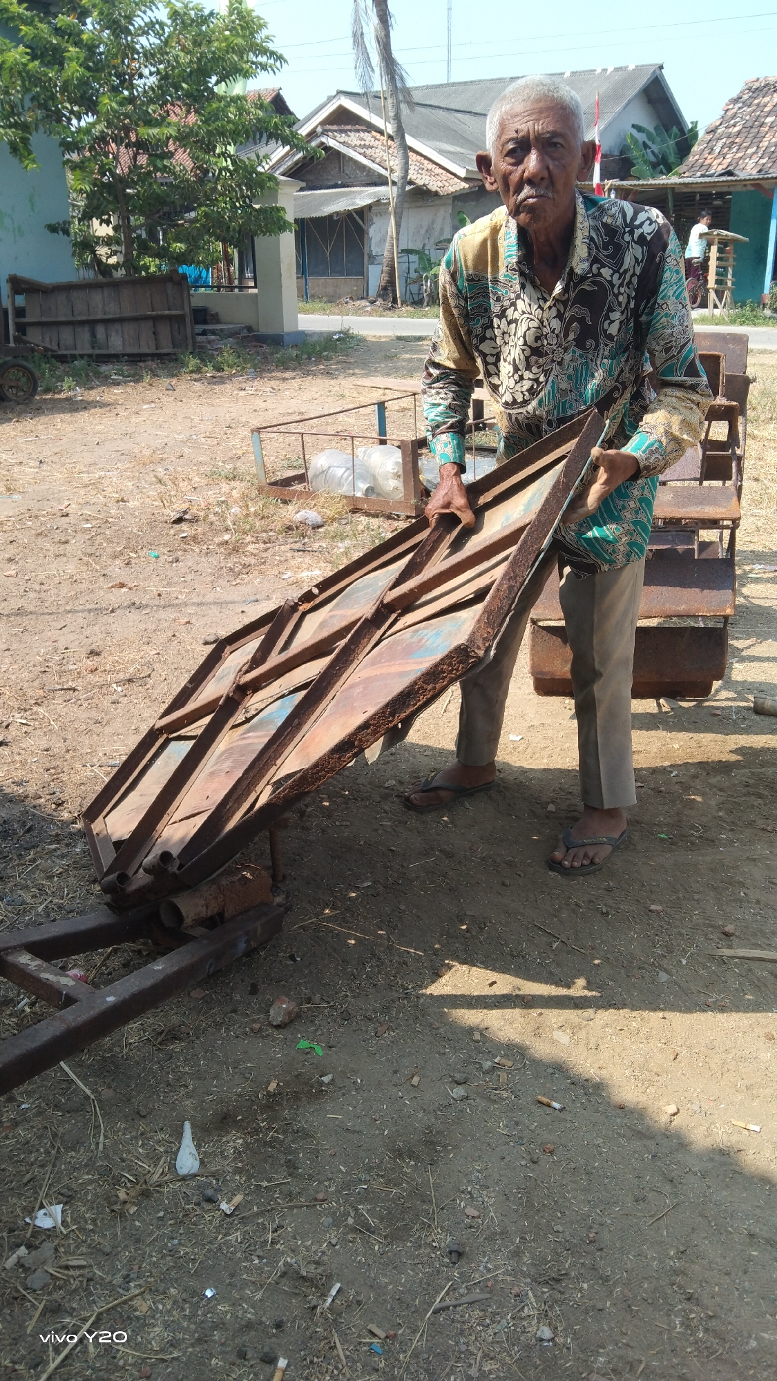 An elderly Indonesian man, possibly of Javanese descent, stands outdoors, holding a rusty metal frame with both hands. He is wearing a long-sleeved batik shirt in shades of green and brown, gray pants, and black flip-flops. His facial features, including his mustache and wrinkles, suggest advanced age. He is looking directly at the camera with a neutral expression.
The scene appears to be a rural or semi-rural area, likely within the city of Karawang, Indonesia, given the context. The ground is unpaved, covered in dirt, dry grass, and scattered debris, including cigarette butts and small stones. In the background, there are several modest buildings with tiled roofs, some appearing to be houses. A lush green tree with dense foliage is visible to the left, and a palm tree stands further back. There are also other discarded metal structures and frames in the vicinity, suggesting a workshop or storage area for old equipment. The lighting indicates it is daytime, with shadows cast on the ground. A watermark in the bottom left corner reads "vivo Y20".